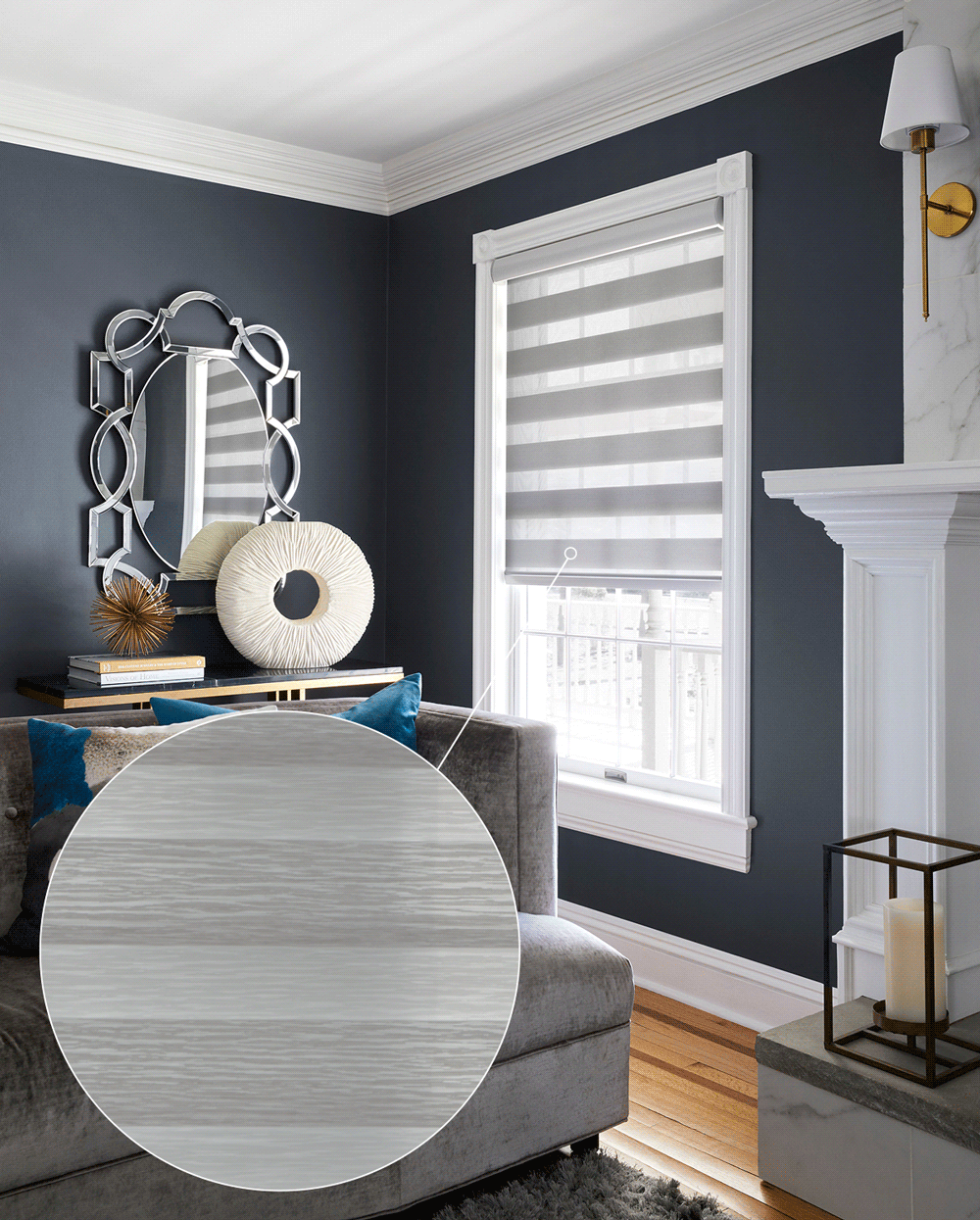 A living room with dark blue walls, a window with gray and white striped blinds, a light gray sofa with blue and beige pillows, a marble fireplace with a gold and white candle holder, a decorative mirror with geometric shapes, and a black console tab