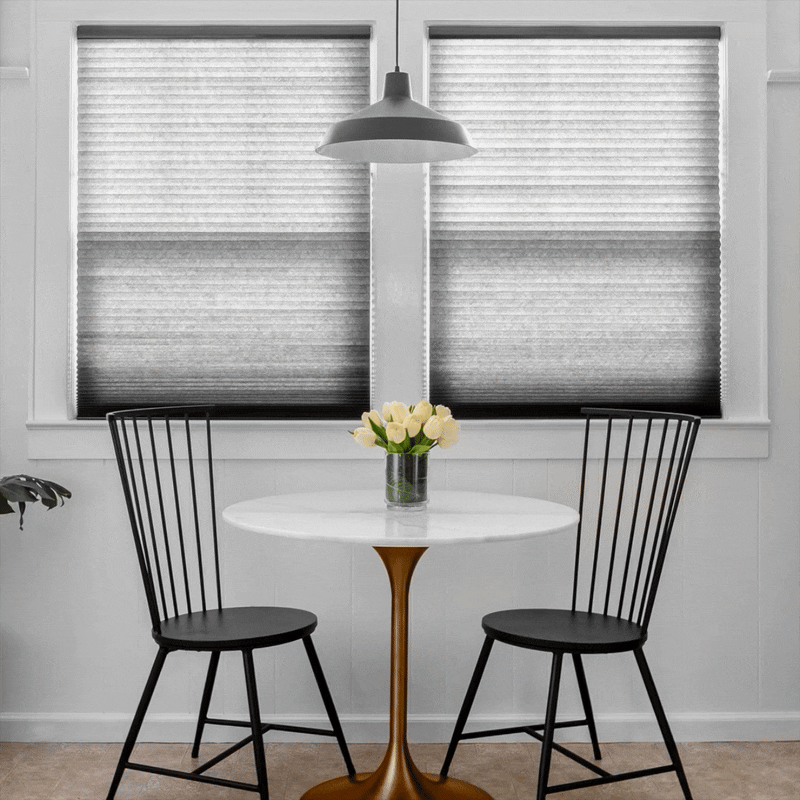 A small dining area with a round white table, two black chairs, a vase with white roses, two window blinds, and a hanging gray pendant light.