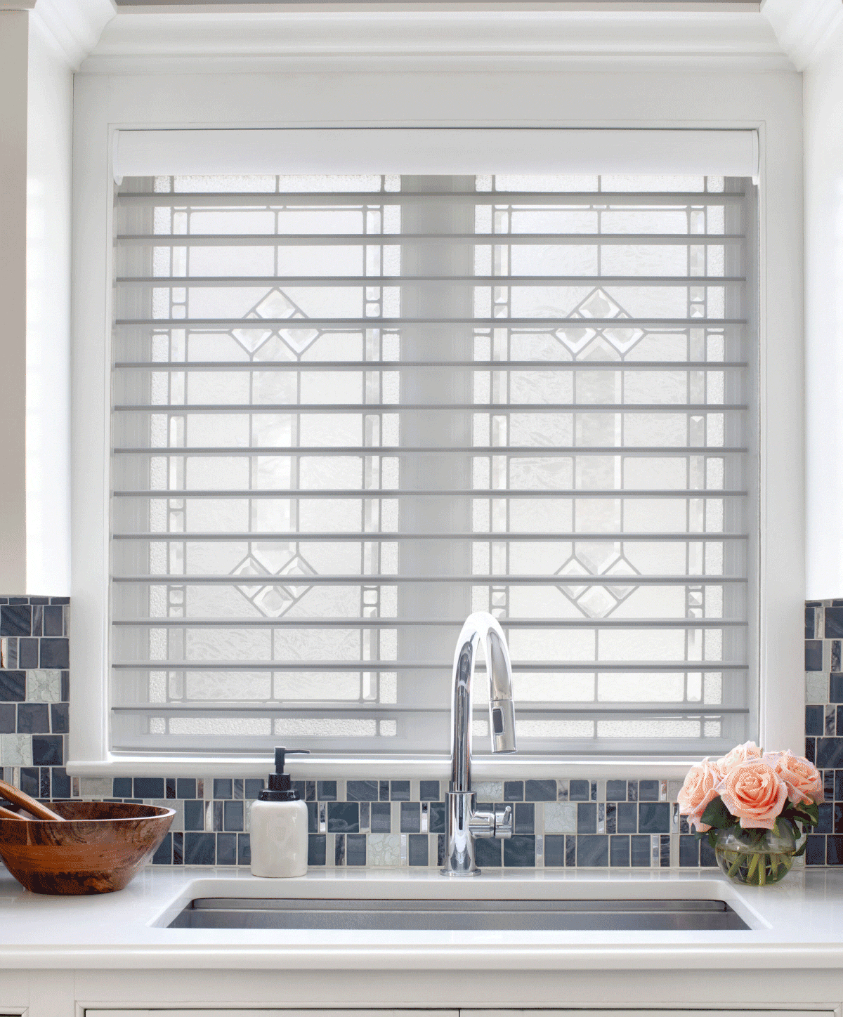 Kitchen sink area with a window covered by blinds, a soap dispenser, a wooden bowl, and a vase of pink roses on the countertop.
