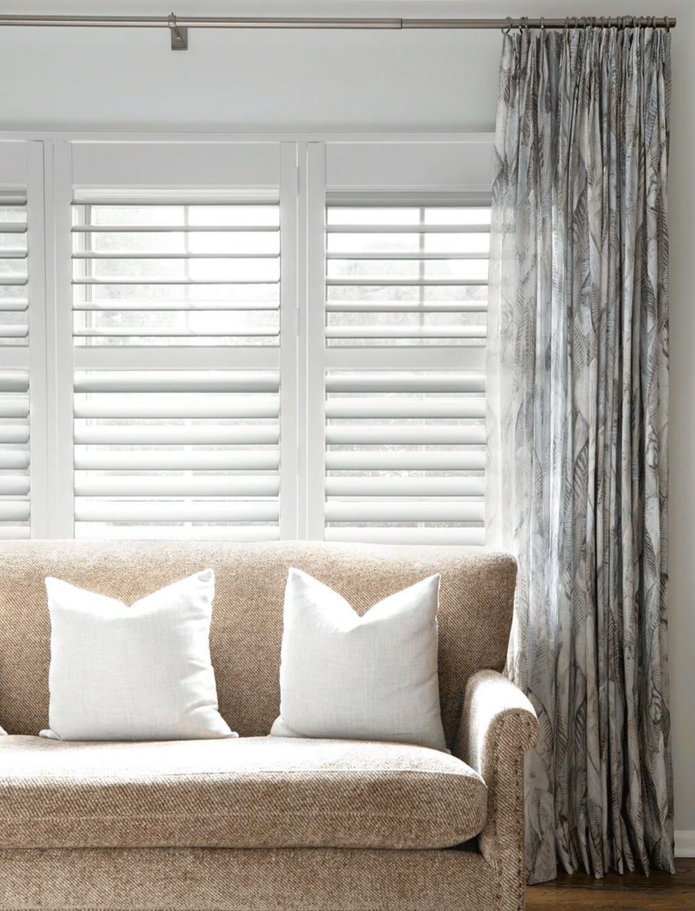 Living room with a beige upholstered sofa, two white pillows, and a window with white blinds and patterned curtains.