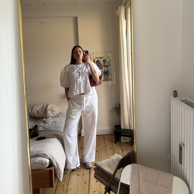 A woman taking a mirror selfie in a bedroom. She is wearing a white, puffy-sleeved blouse and matching white wide-leg pants. The room has a bed with beige bedding, a small black side table with a plant, and a large mirror with a gold frame. Natural light is coming through a window with white curtains.