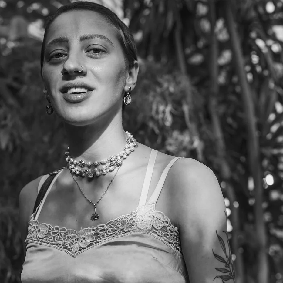 A woman with short hair, wearing jewelry and lace detail top, standing outdoors with trees in the background.