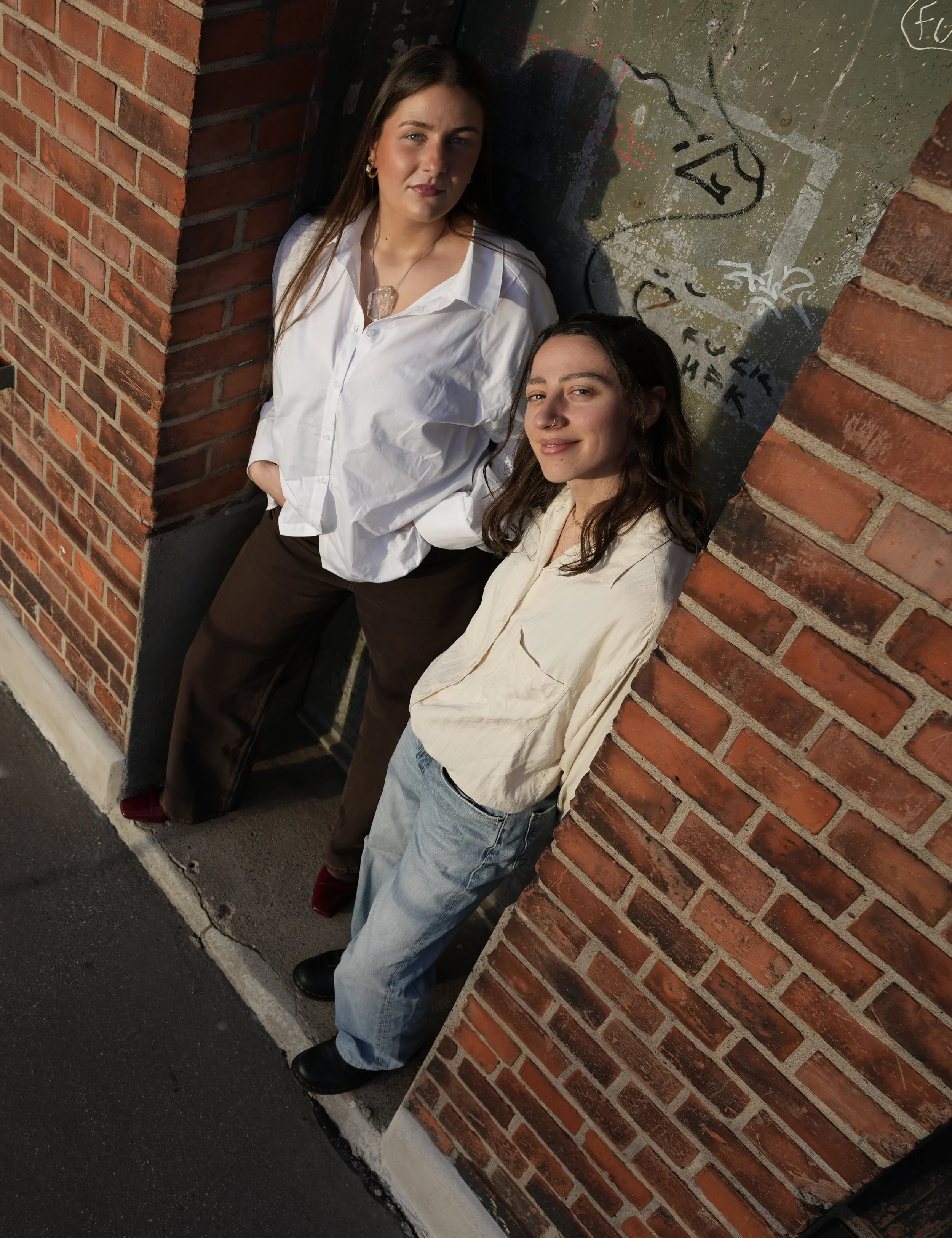 Two young women leaning against a brick corner on a sidewalk with graffiti on a concrete wall behind them. One woman has long brown hair, fair skin, wearing a white shirt and brown pants. The other woman has long dark hair, tanned skin, wearing a cream-colored jacket and light-colored jeans.