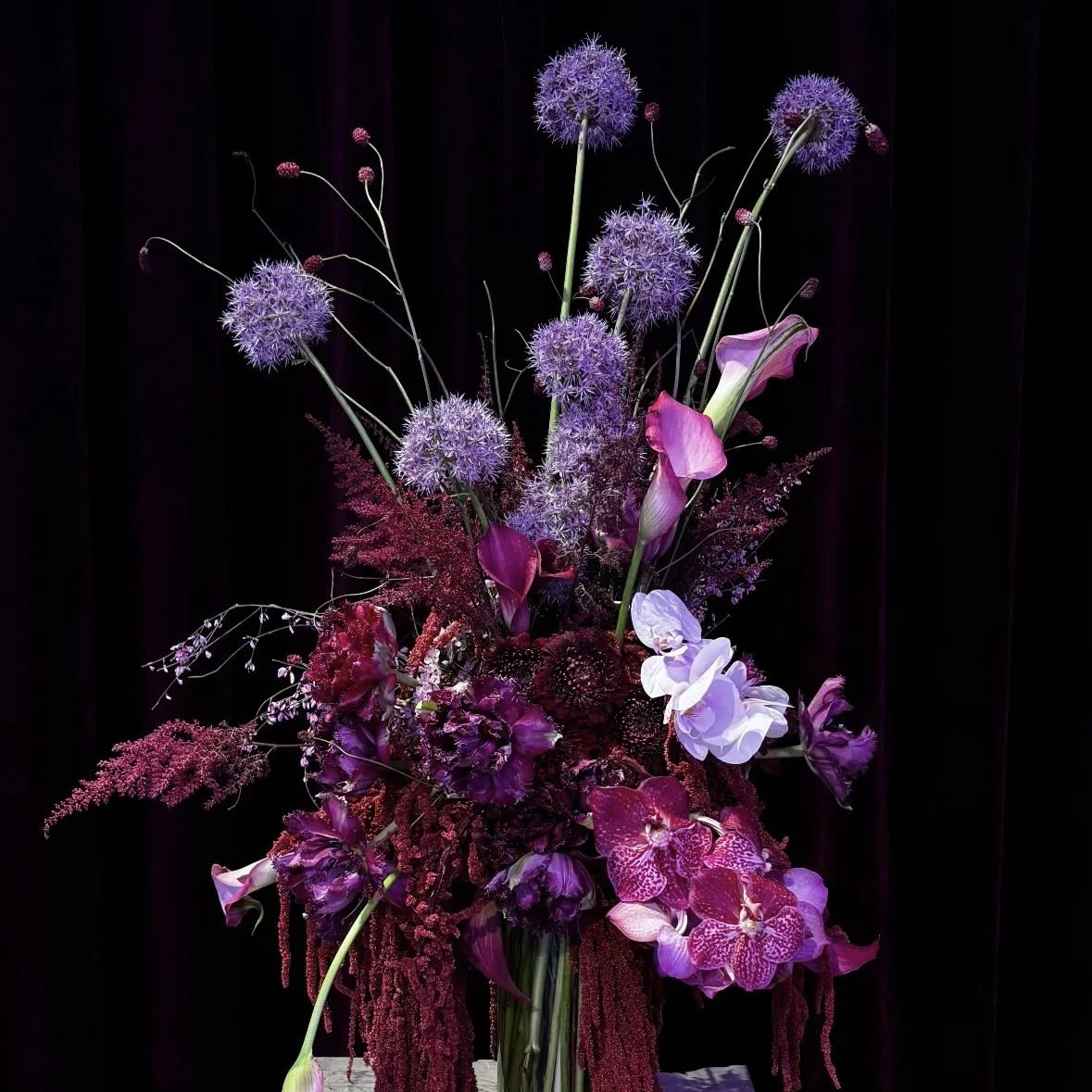 A vibrant bouquet of purple, pink, and white flowers, including alliums and orchids, arranged in a glass vase against a dark background.