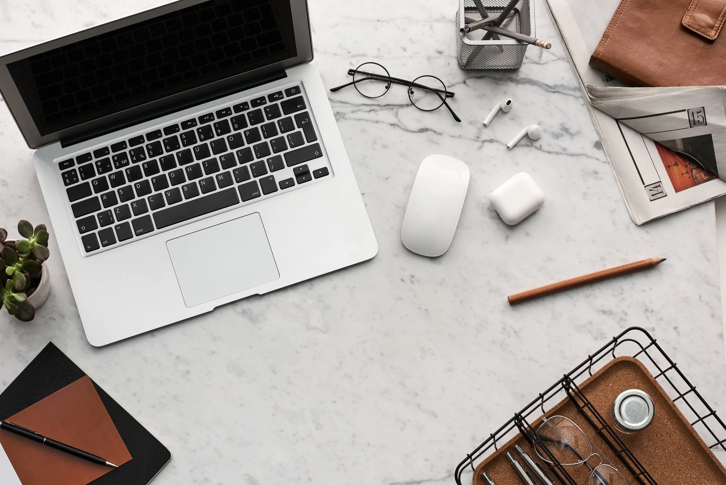 A workspace with a laptop, glasses, wireless earbuds, a mouse, a journal, a pencil, a magazine, a small plant, and office supplies on a white marble table.