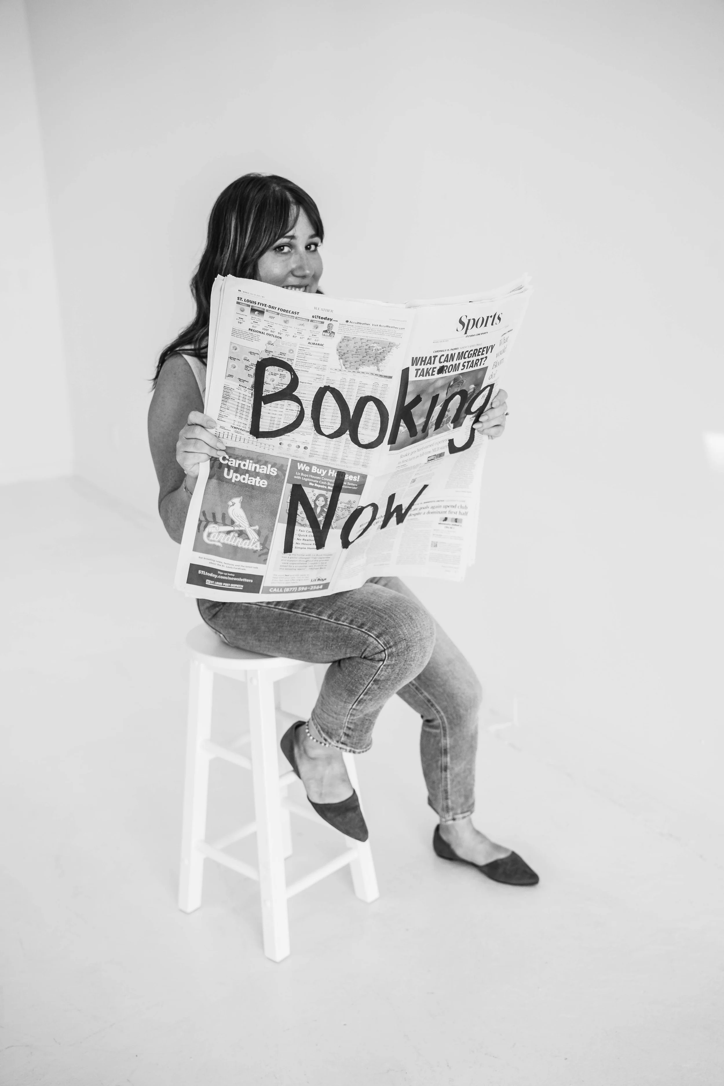 A woman sitting on a white stool, holding a newspaper with 'Booking Now' written across it in large letters.