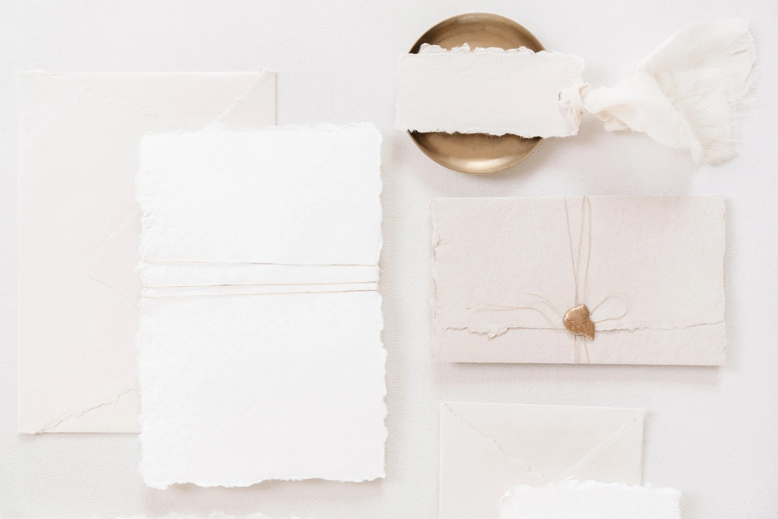 Flat lay of blank white and cream-colored textured paper, some torn, along with a gold sealed envelope, a gold dish, and a piece of white tissue paper on a white background.