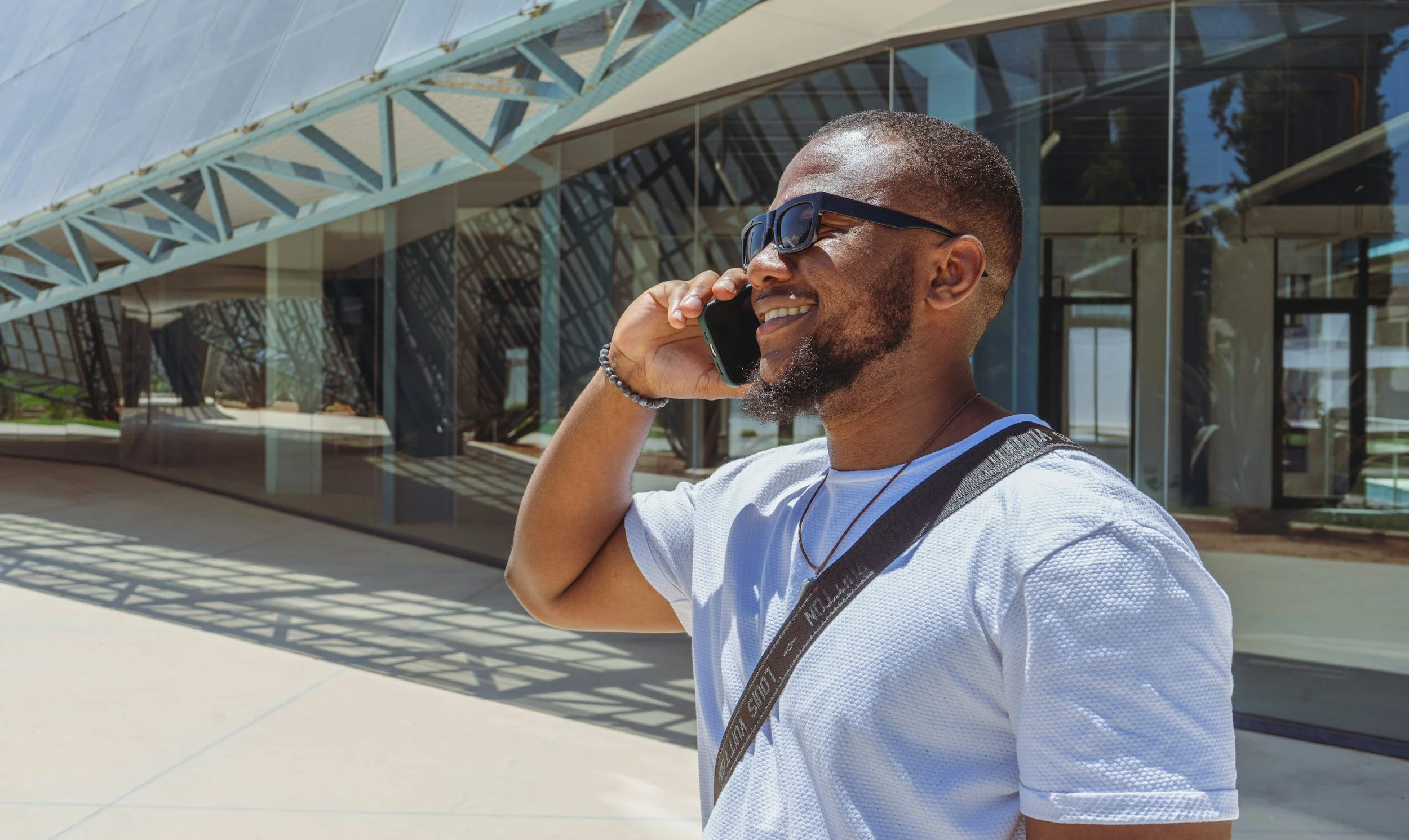 A man with a beard and sunglasses talking on his phone outside a modern building with glass walls and metal framework.