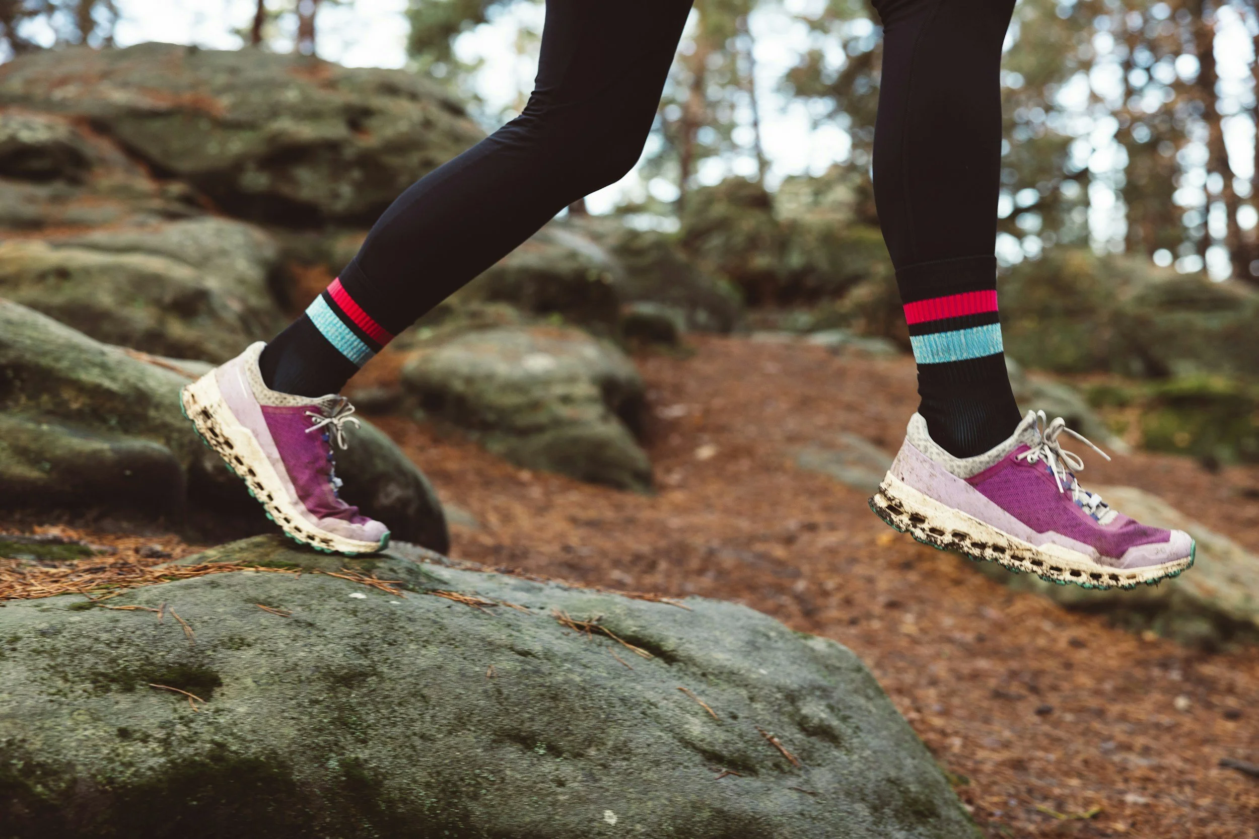 Person wearing pink running shoes, black leggings with colorful stripes, and black socks, climbing over a rocky trail in a forested area.