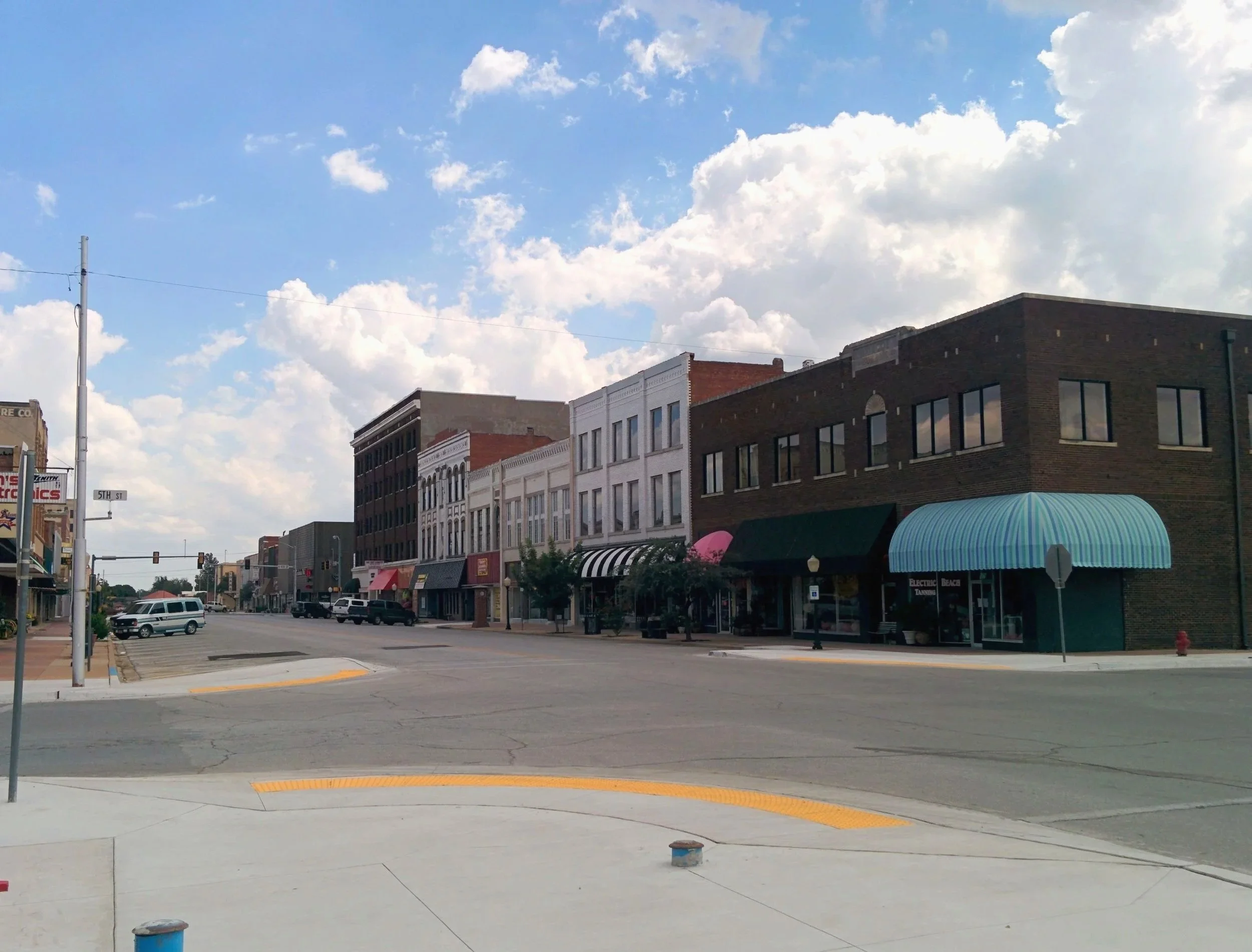 Empty Main Street in a small town with old buildings, storefronts with awnings, parked cars, sidewalk, and blue sky with clouds.
