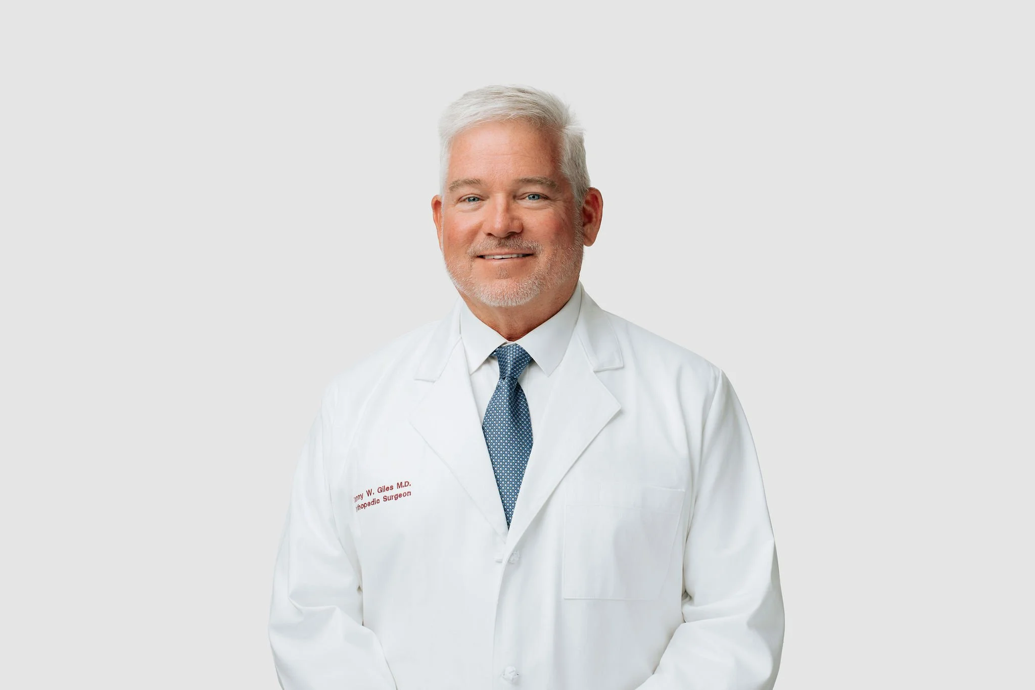 A middle-aged man with gray hair and a beard, wearing a white lab coat and a blue tie, smiling at the camera against a plain light gray background.
