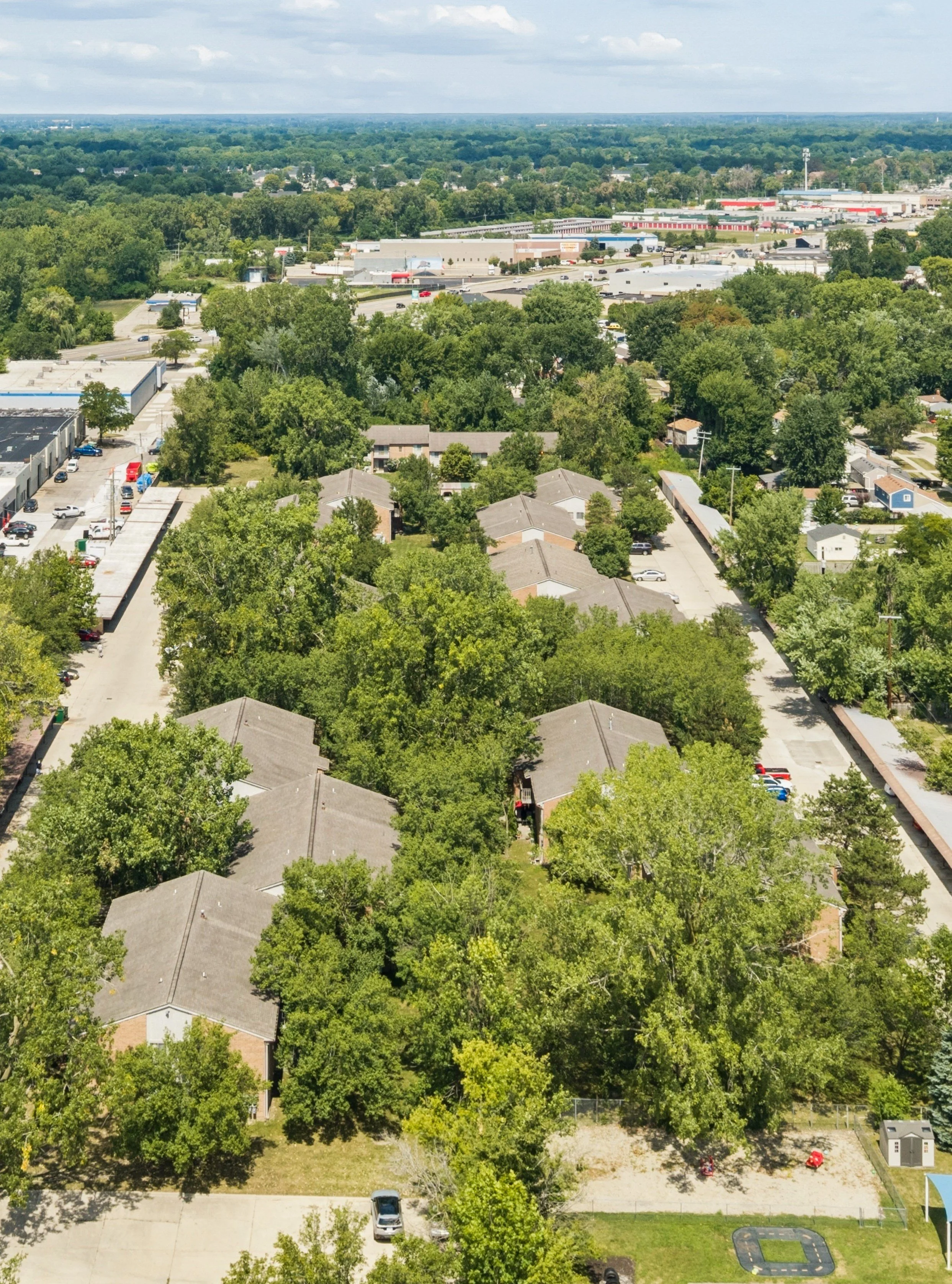 An aerial view of a residential neighborhood with houses, trees, parking areas, and green lawns under a partly cloudy sky.