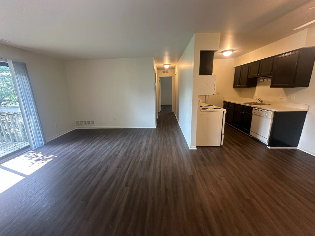 Empty living room and kitchen area in an apartment with hardwood floors, white walls, sliding glass door leading to balcony, and black kitchen cabinets.