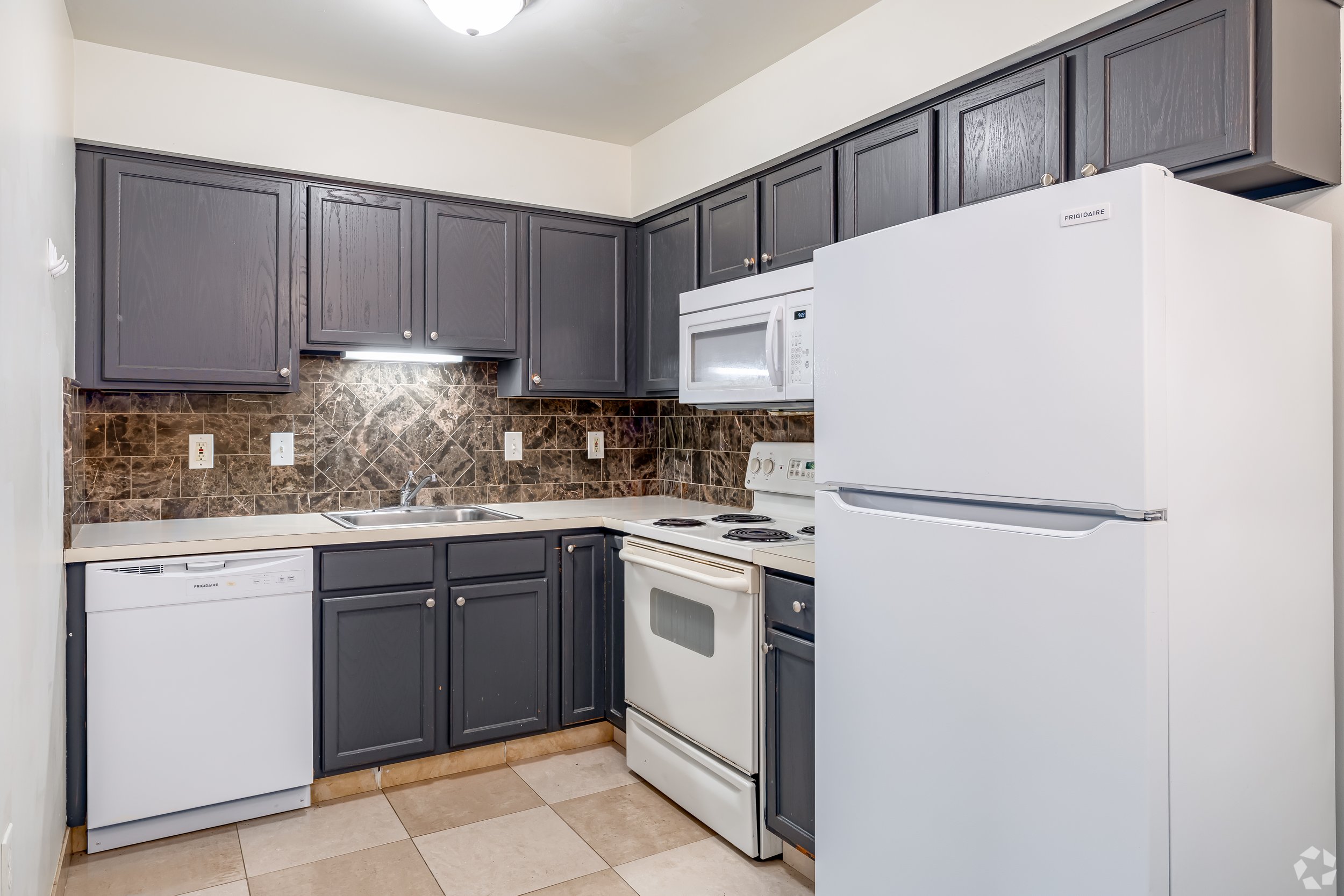 Kitchen with dark gray cabinets, a white refrigerator, white microwave, white stove, dishwasher, and a brown marble backsplash.