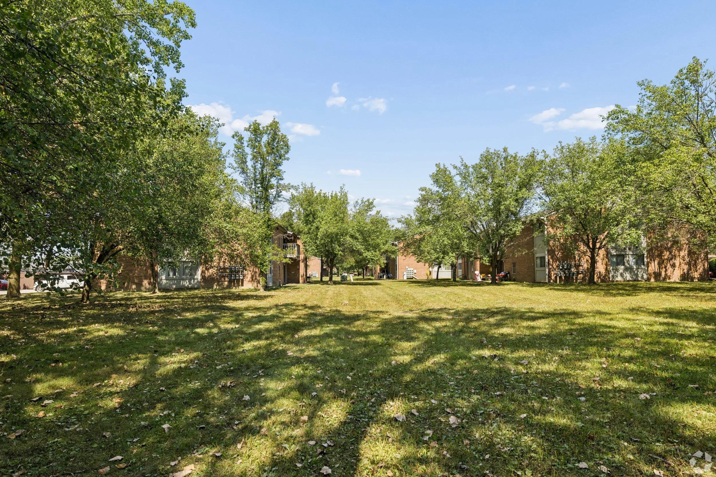 A residential apartment complex with brick buildings, surrounded by blooming white trees on a grassy lawn, under a partly cloudy sky.
