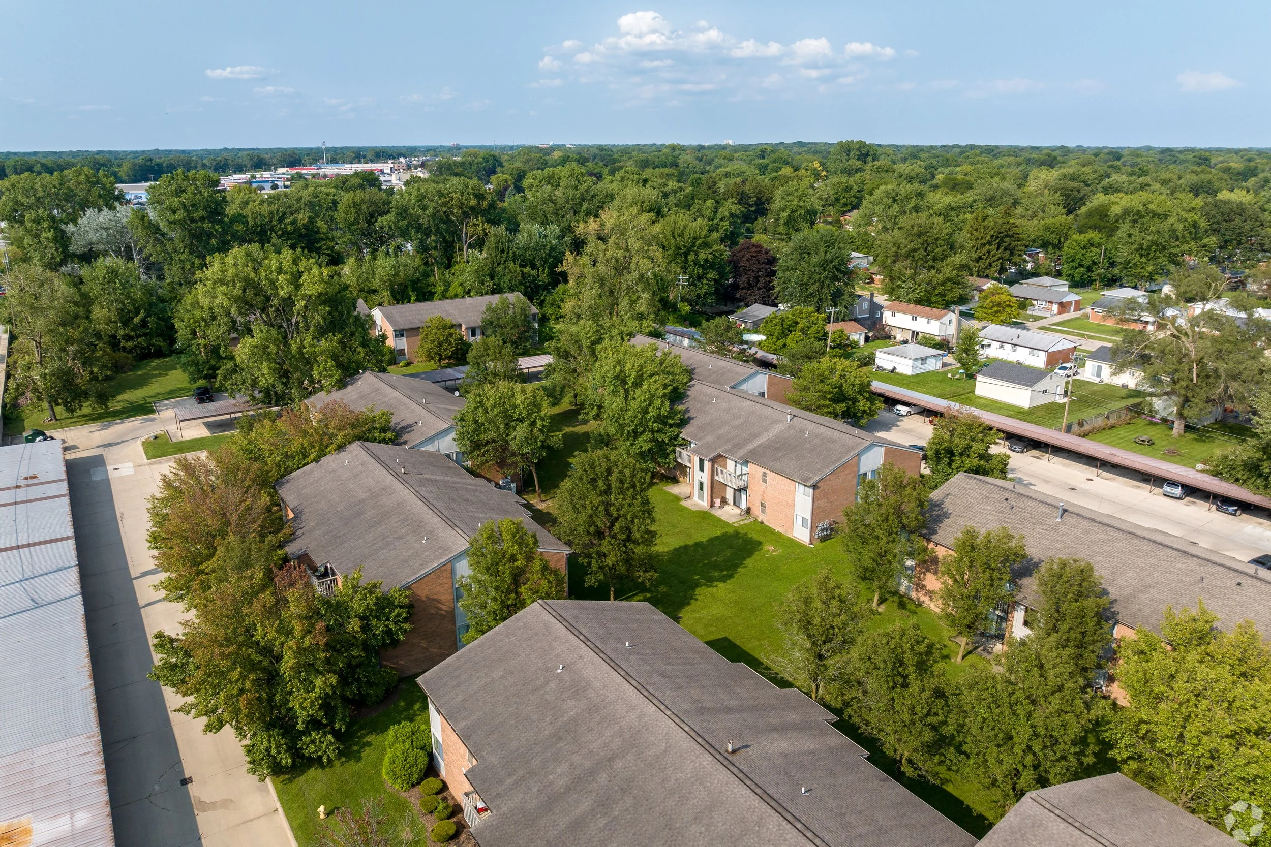 An aerial view of a residential neighborhood with houses, trees, parking areas, and green lawns under a partly cloudy sky.