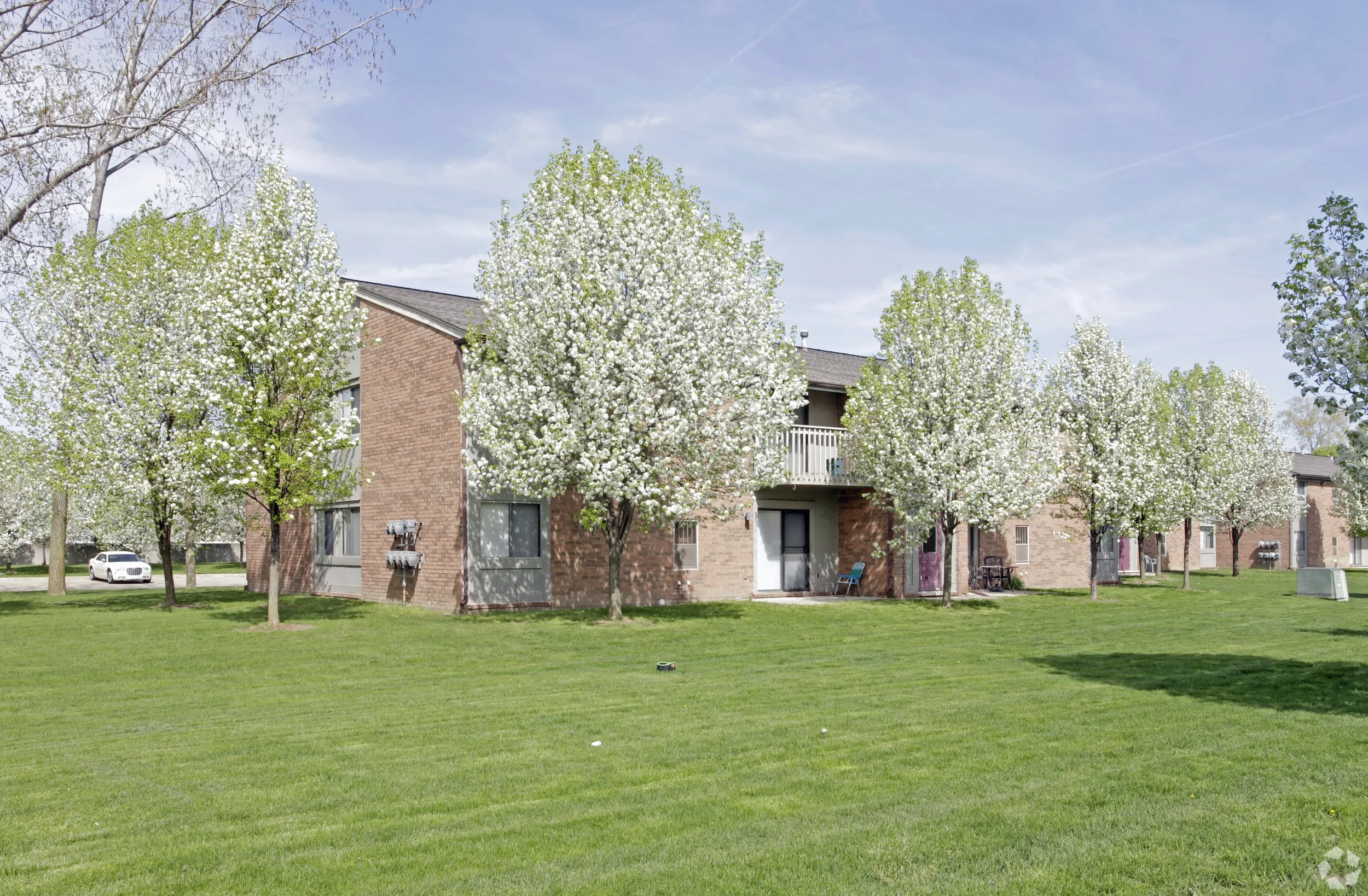A grassy area with several blooming trees with white flowers in front of a two-story brick apartment building, under a partly cloudy sky.