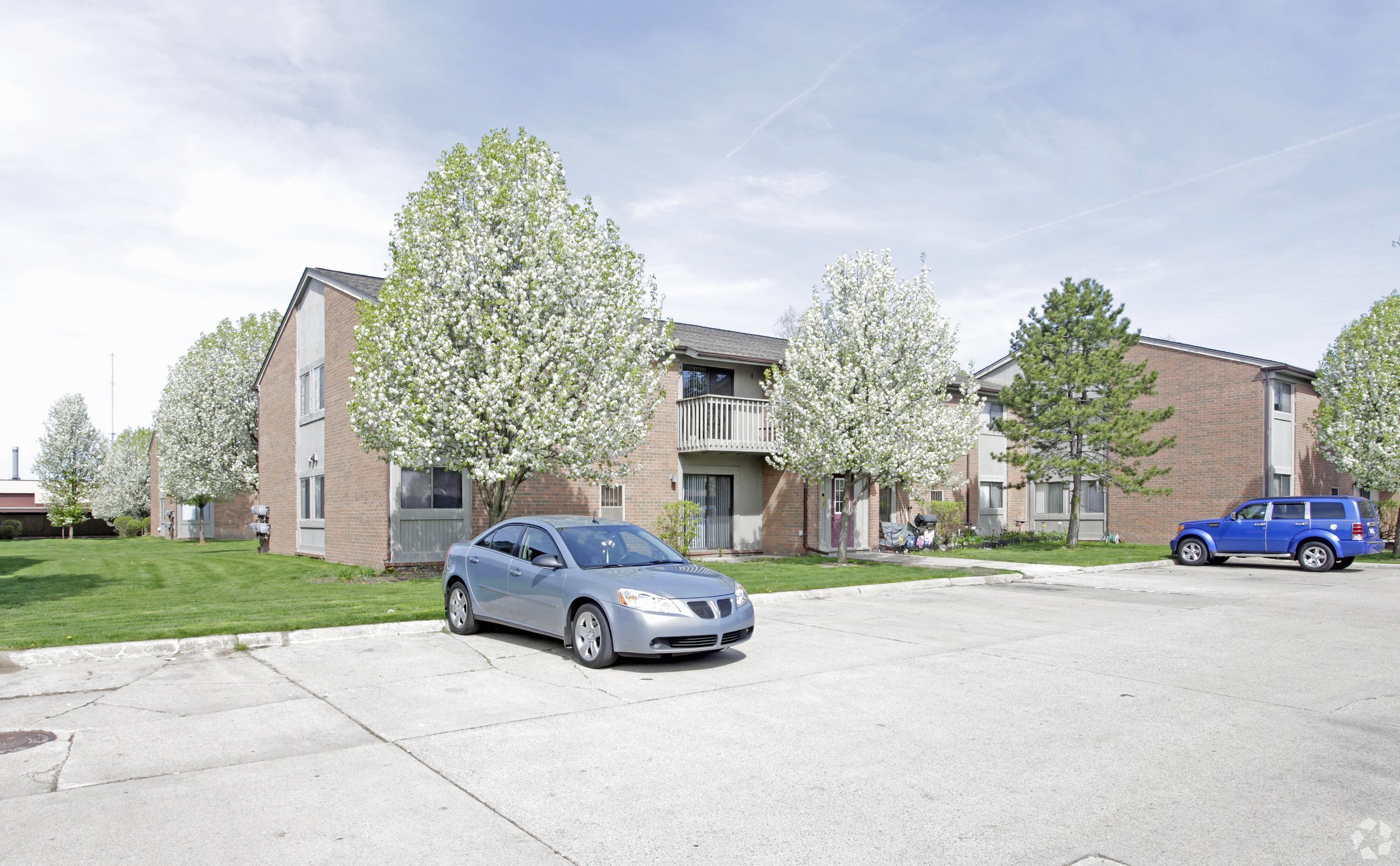 An apartment complex with brick buildings, green lawns, and blooming trees in spring. Two cars, a silver sedan and a blue SUV, are parked in the lot.