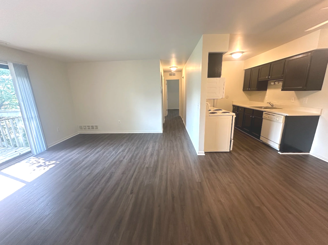 Empty living room with sliding glass door leading to a balcony, and an open kitchen with black cabinets and white appliances.