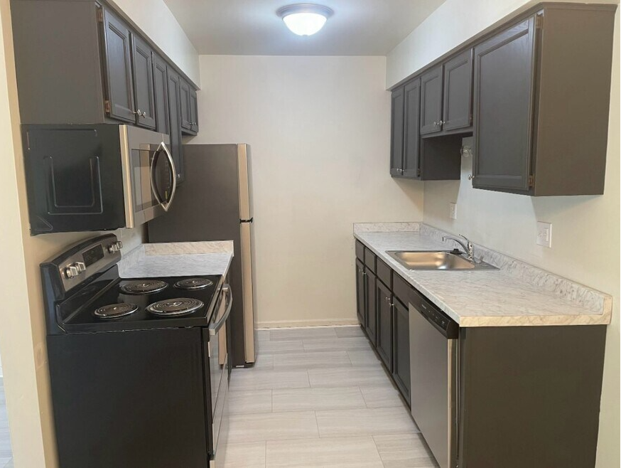 A small kitchen with dark gray cabinets, a stainless steel refrigerator, microwave, and dishwasher, a black electric stove, and a marble countertop with a sink.