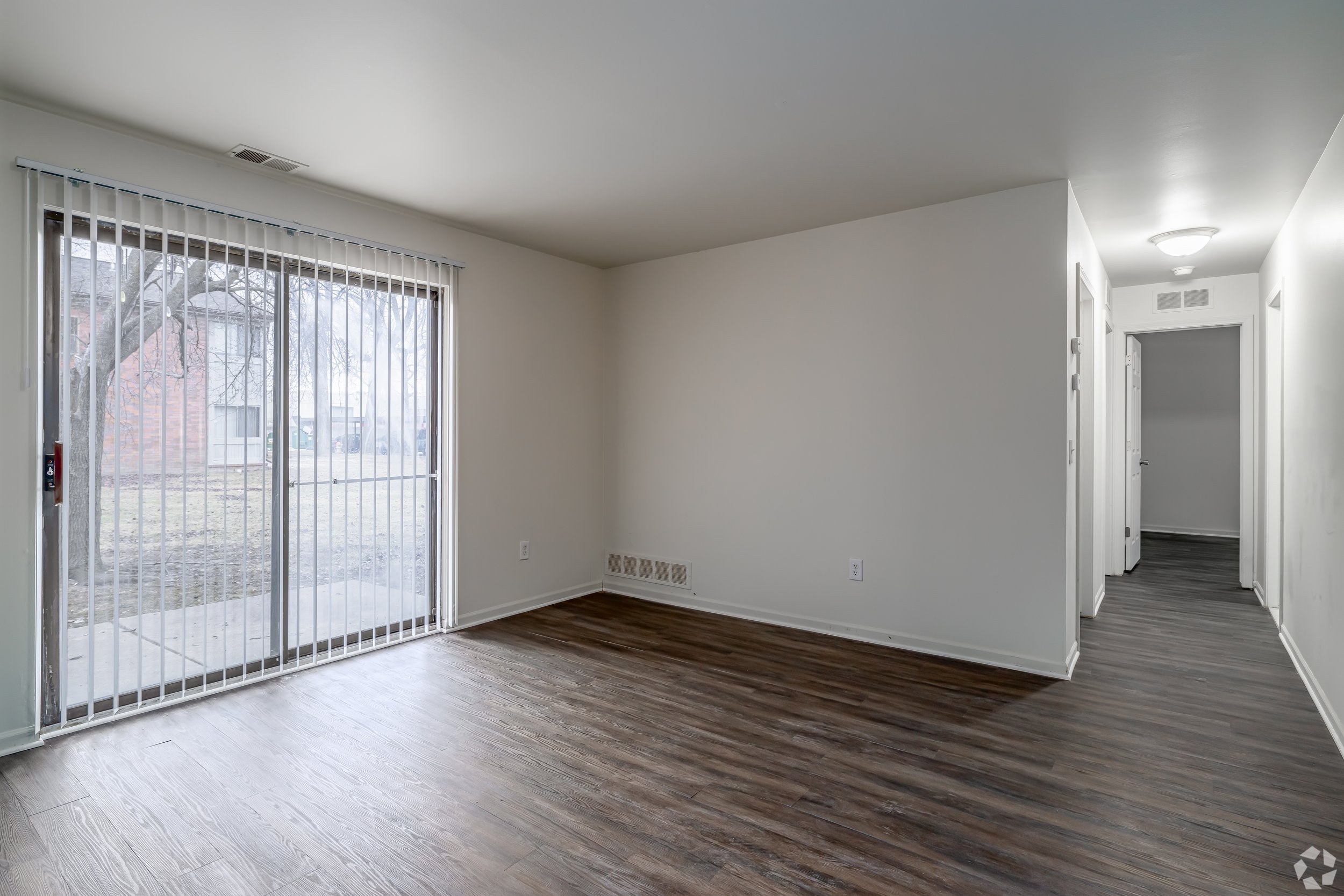 Empty living room with sliding glass door, white walls, and wooden floor, near a hallway.