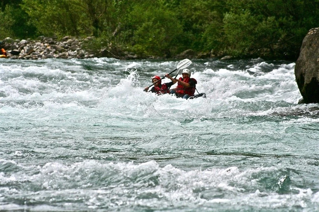Two people white water rafting on a river with green trees and rocks in the background.