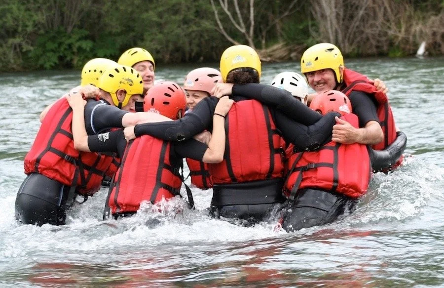 Group of people wearing helmets and life jackets, participating in a river rafting activity, embracing each other in a huddle on the water.