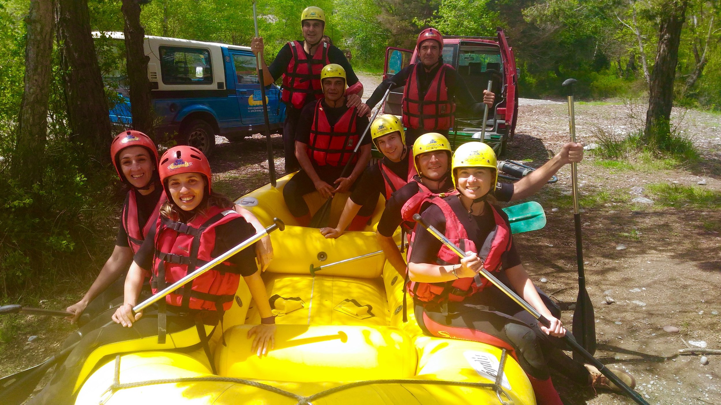 Group of people in life jackets and helmets preparing for white-water rafting on a yellow raft in a wooded outdoor area.