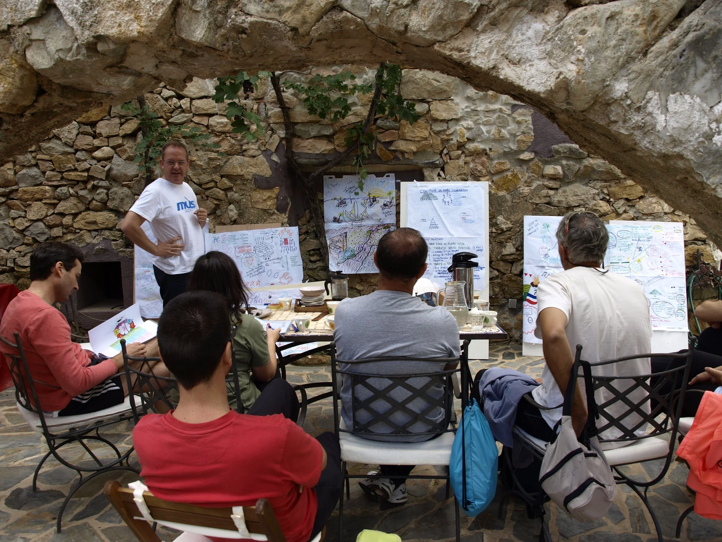 A group of people gathered in a courtyard with stone walls and an arch ceiling, attending a presentation. A man in a white T-shirt stands near a whiteboard with drawings and notes, addressing the audience. Several posters with drawings and notes are displayed on the stone wall.