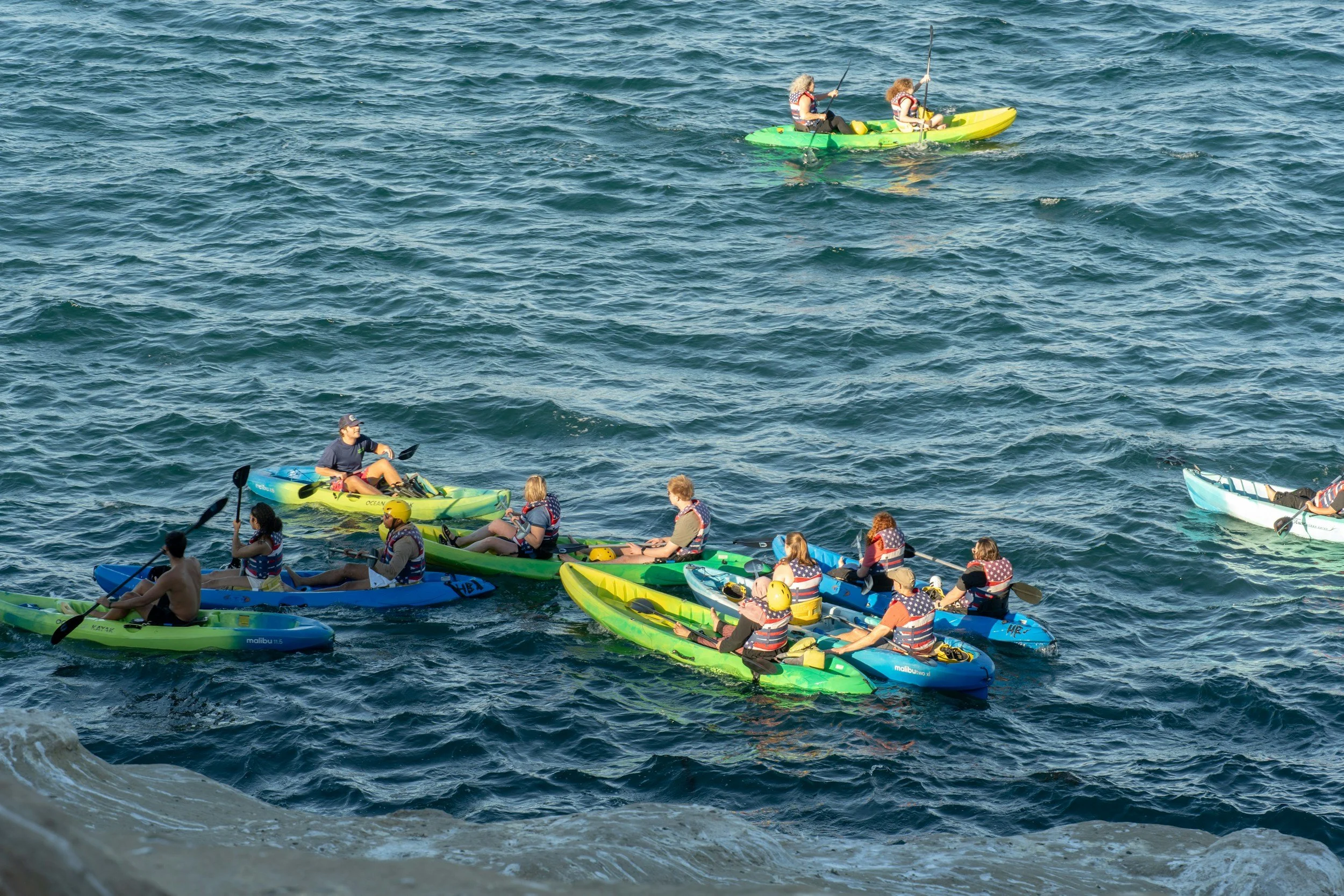 Multiple people kayaking on a large body of water, some wearing life jackets with American flag patterns, with a view from inside a small rocky opening.
