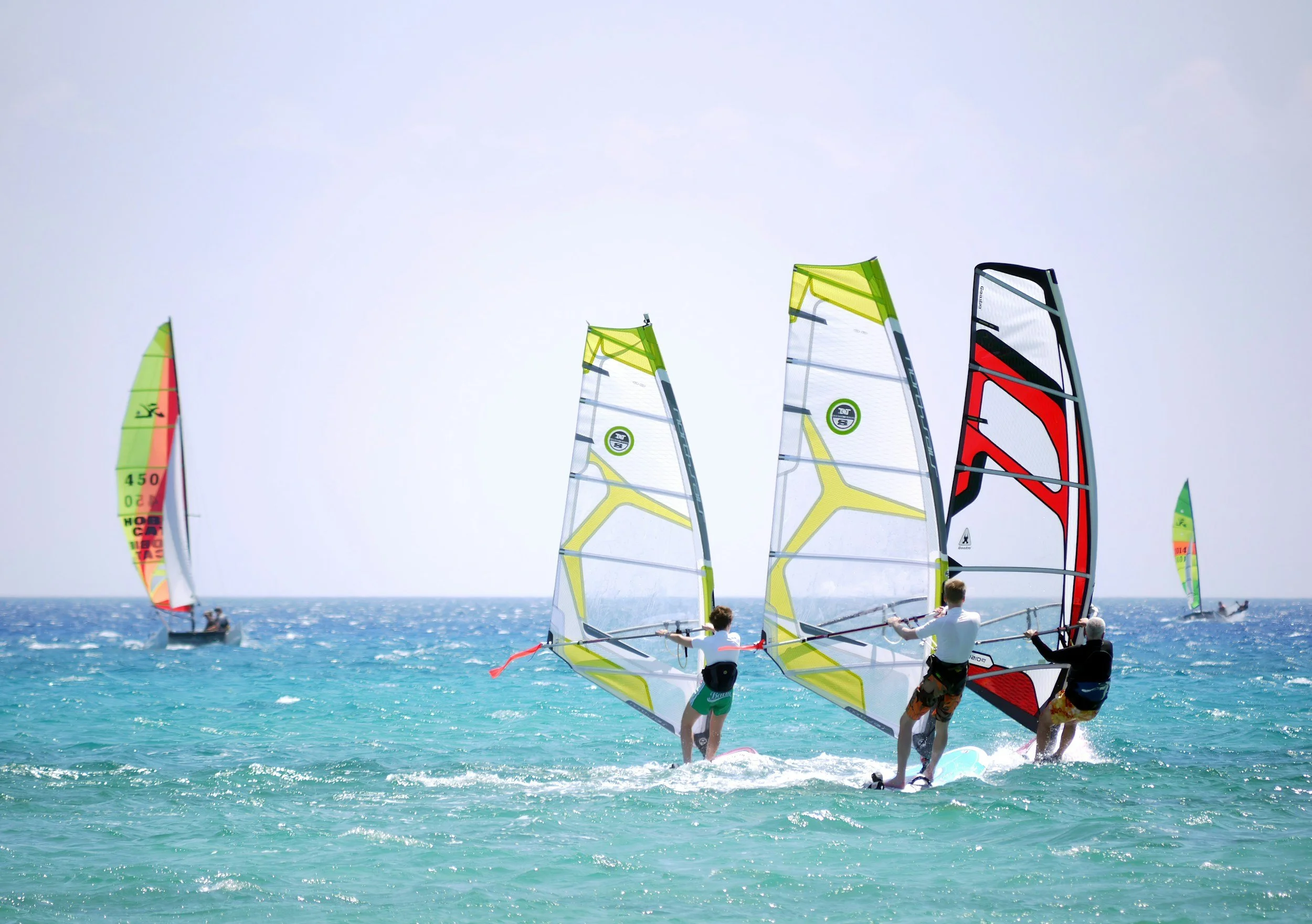 Group of windsurfers riding on the ocean with colorful sails, some standing on their boards, under a clear sky.