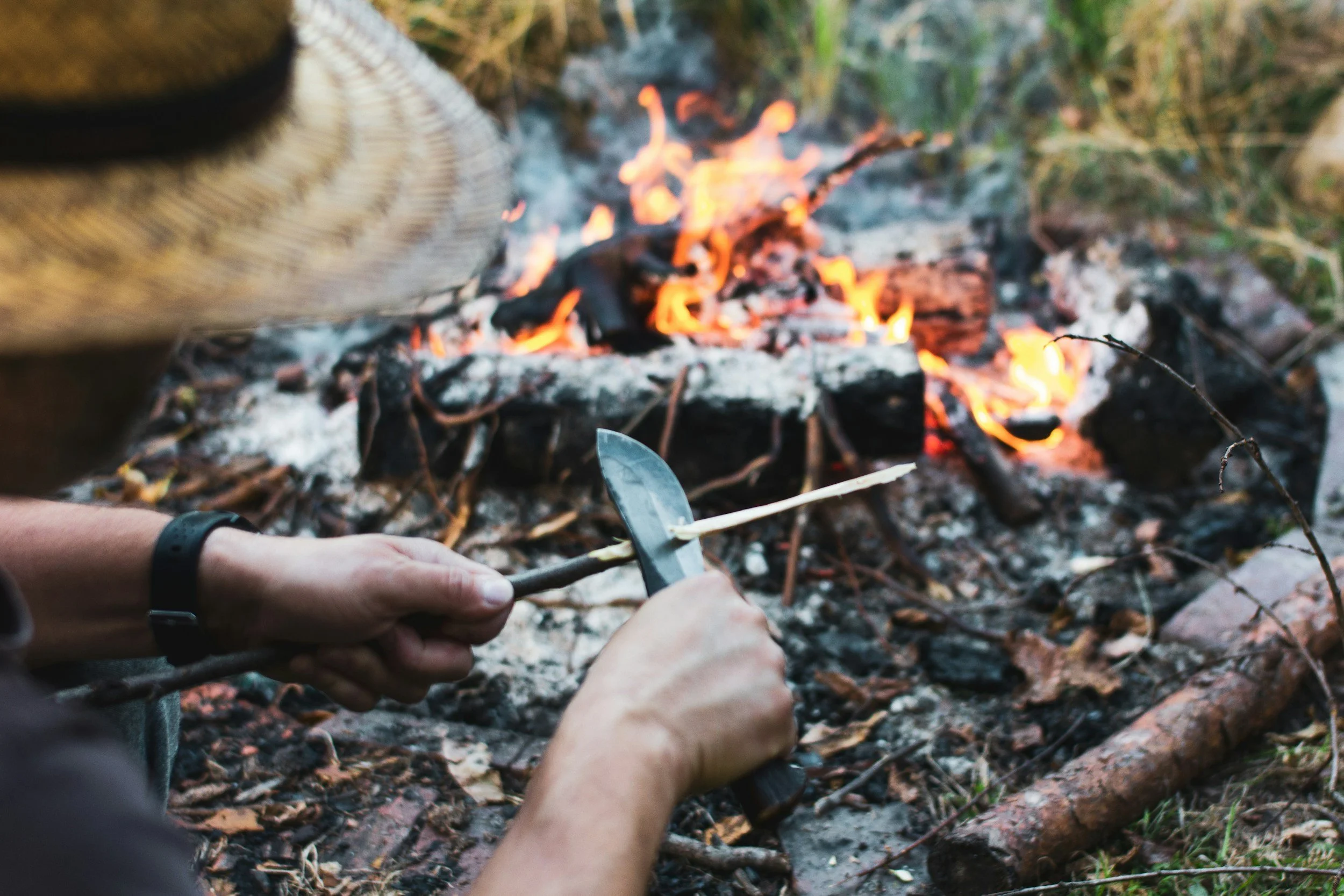 Person starting a campfire outdoors, holding a stick and a small knife, with a blaze of flames and smoke in the background.