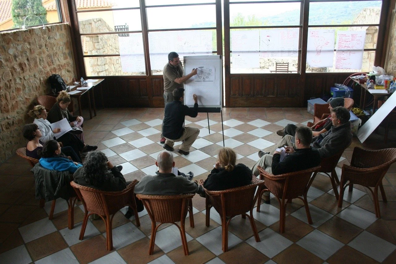 A man giving a presentation on a whiteboard in a room with large windows and several audience members seated in a semi-circle around him.
