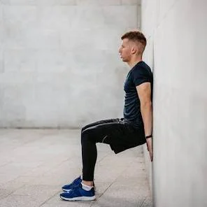 A man wearing a navy t-shirt and black pants performs a wall squat exercise against a plain white wall.