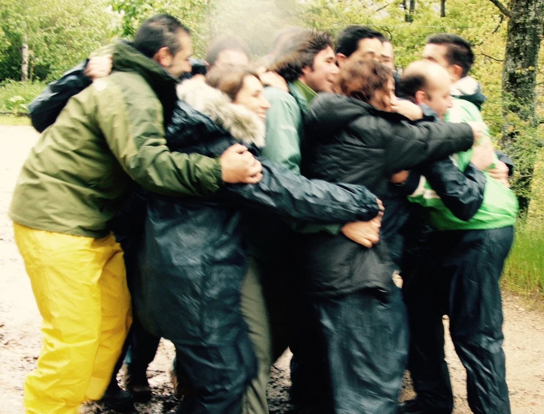 Group of people standing outdoors in a close huddle, embracing each other and smiling, with trees in the background.