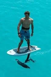 A young man standing on a paddleboard in the water, holding a kayak paddle, with a seal swimming nearby.