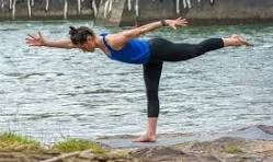 Person practicing yoga on a riverbank, balancing on one leg with arms extended