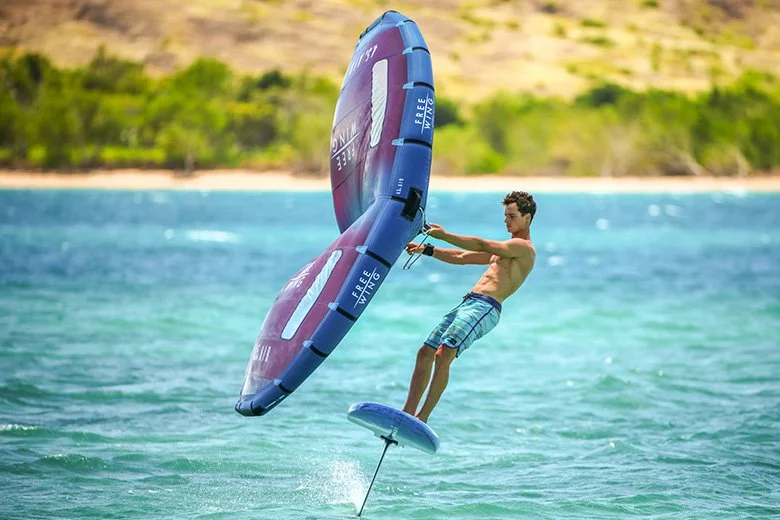 A man is hanging onto a large water-powered kite while riding a hydrofoil in the water, with land and trees in the background.