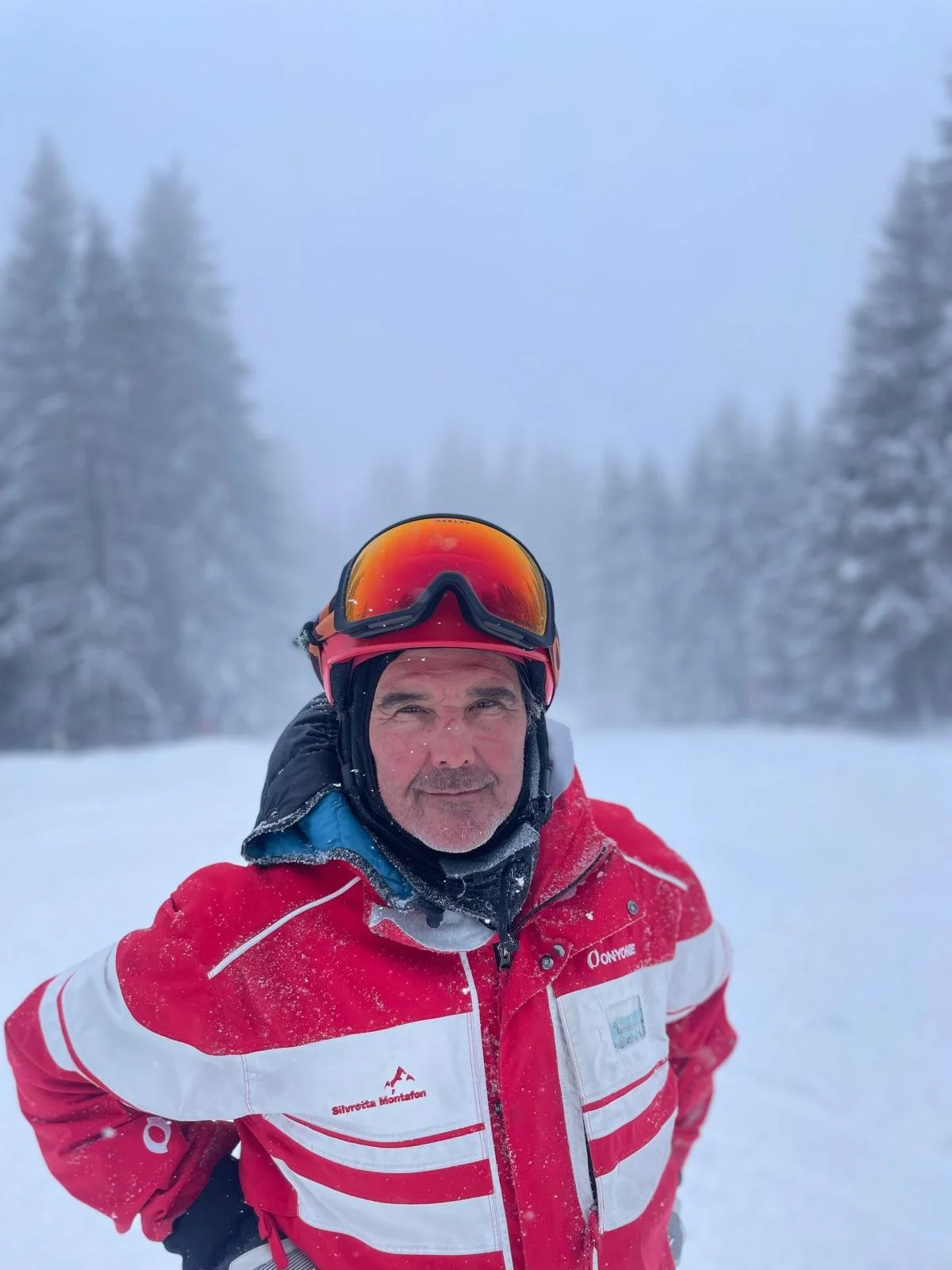 Man wearing red and white winter jacket, black helmet, and ski goggles standing outdoors in snowy landscape, with snow-covered trees in the background.