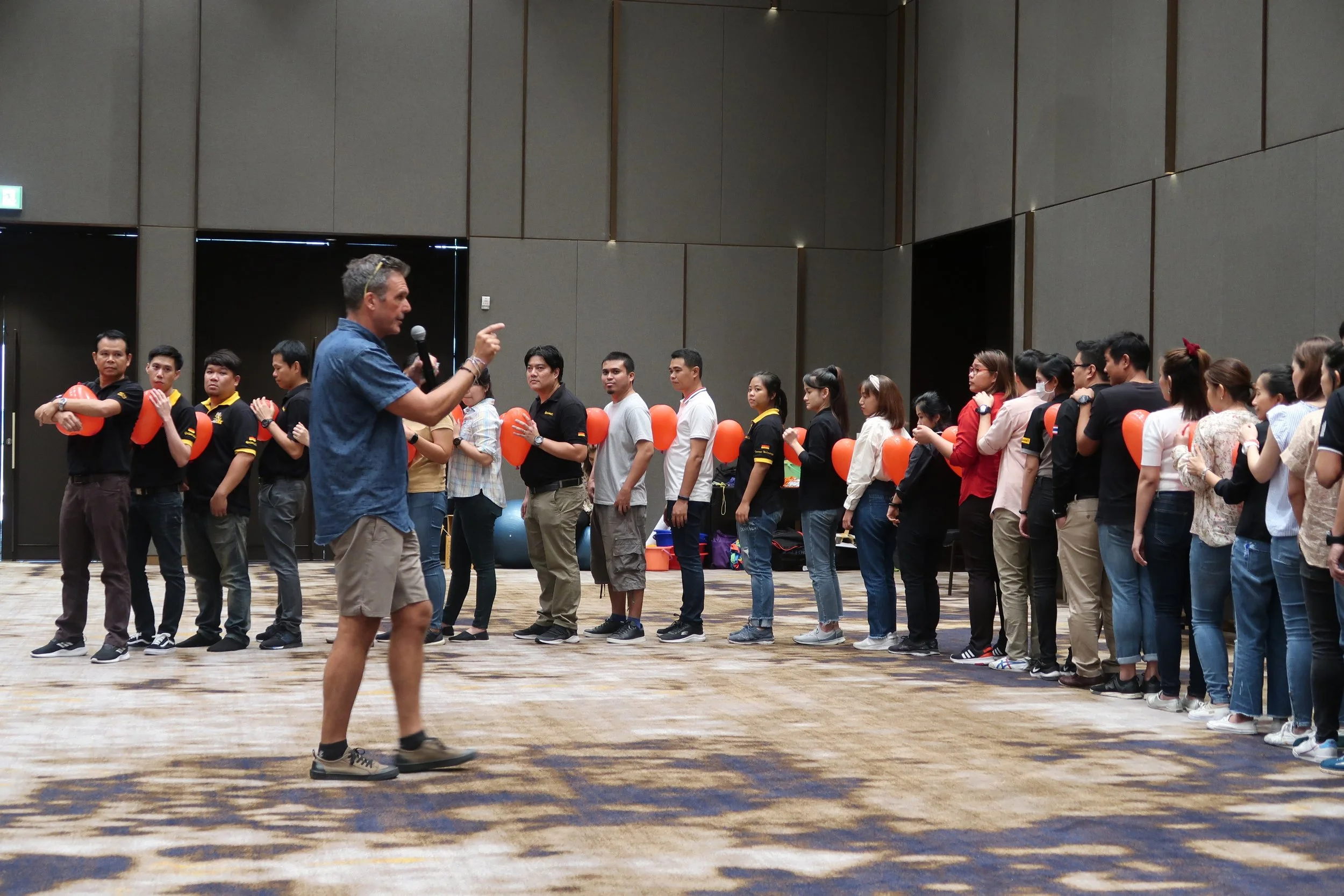A group of people standing in a line, some holding red balloons, with a man in a blue shirt and khaki shorts speaking to them at an indoor event.