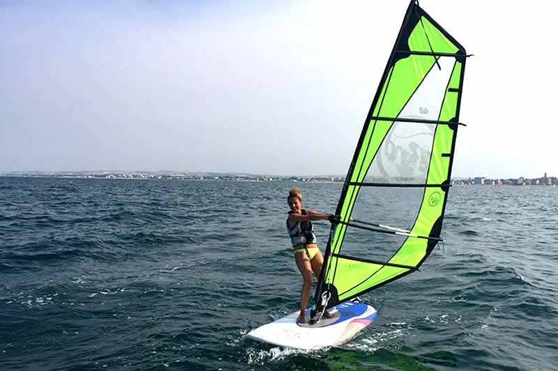 A woman riding a windsurfing board on the water with a bright green sail, wearing a life vest and shorts.