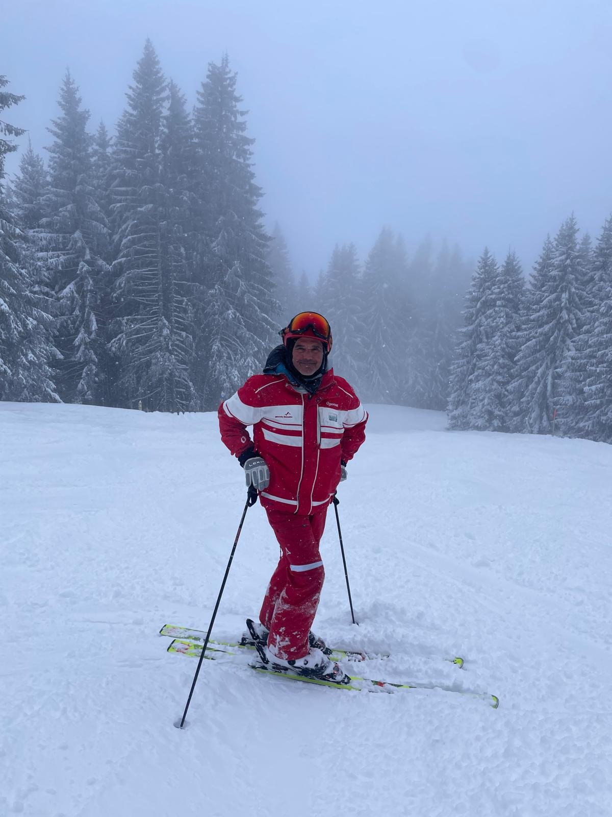 A man dressed in red and white ski gear, wearing a helmet and goggles, standing on skis in a snowy forest with tall pine trees and foggy weather.