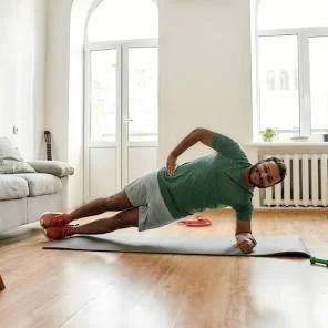 A woman doing a side plank exercise on a yoga mat in a bright living room with large windows.