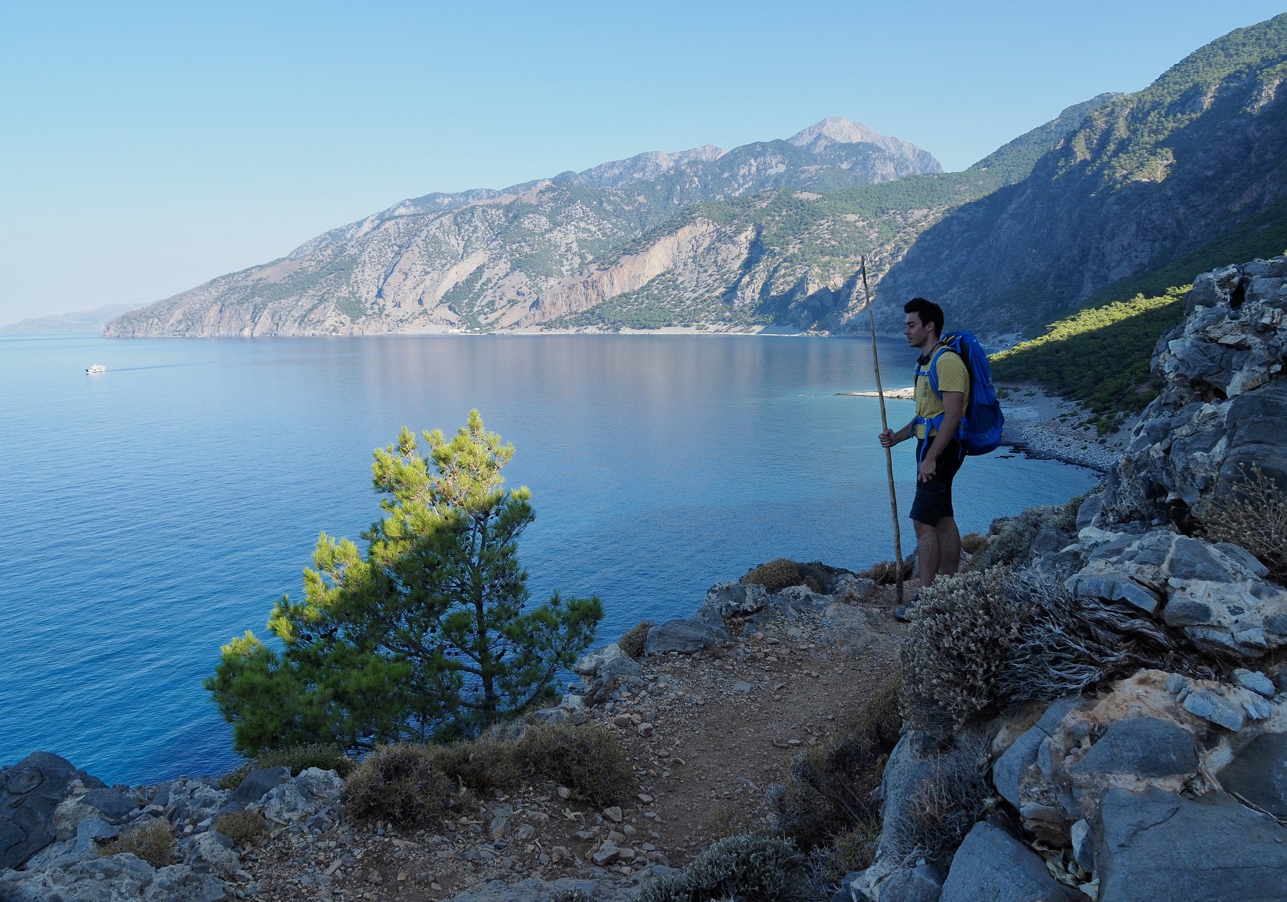 A hiker in a yellow shirt with a blue backpack standing on rocky terrain near a large body of water, with green mountains and a clear blue sky in the background.