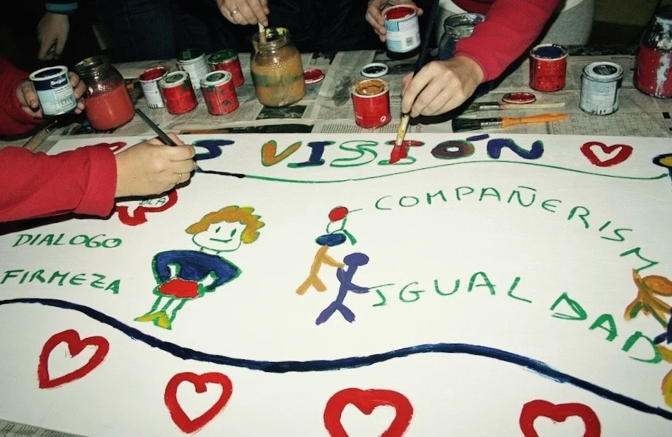 People painting a large banner with colorful text and drawings, using various paint cans and brushes, surrounded by heart symbols.