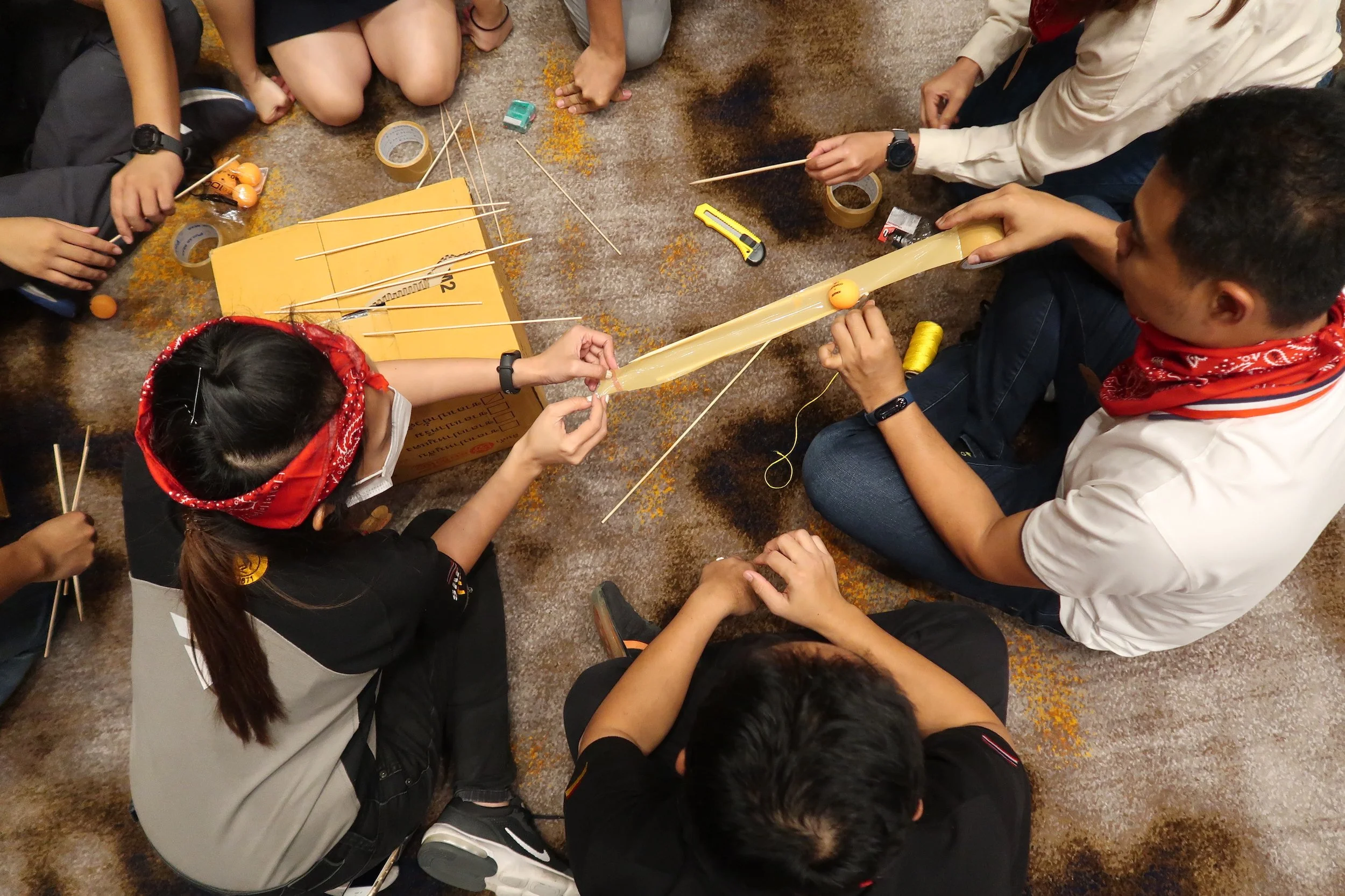 Group of people sitting on the floor making a homemade catapult using rubber bands, sticks, and a bottle. They are assembling the catapult with orange ping pong balls as projectiles. The scene appears to be in an indoor setting with a carpeted floor.