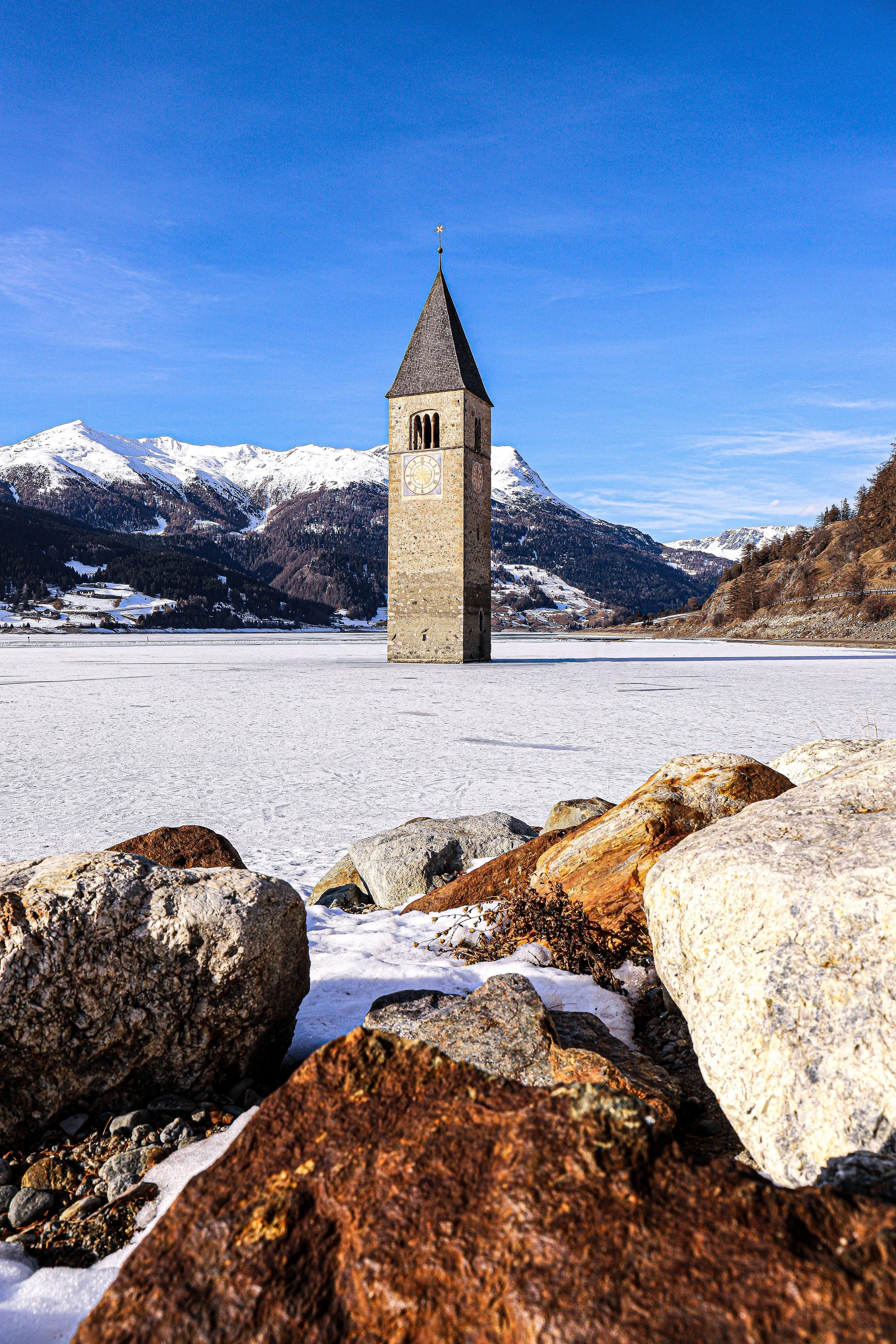 Campanile di pietra in mezzo a un lago ghiacciato con montagne innevate sullo sfondo e rocce sul primo piano.