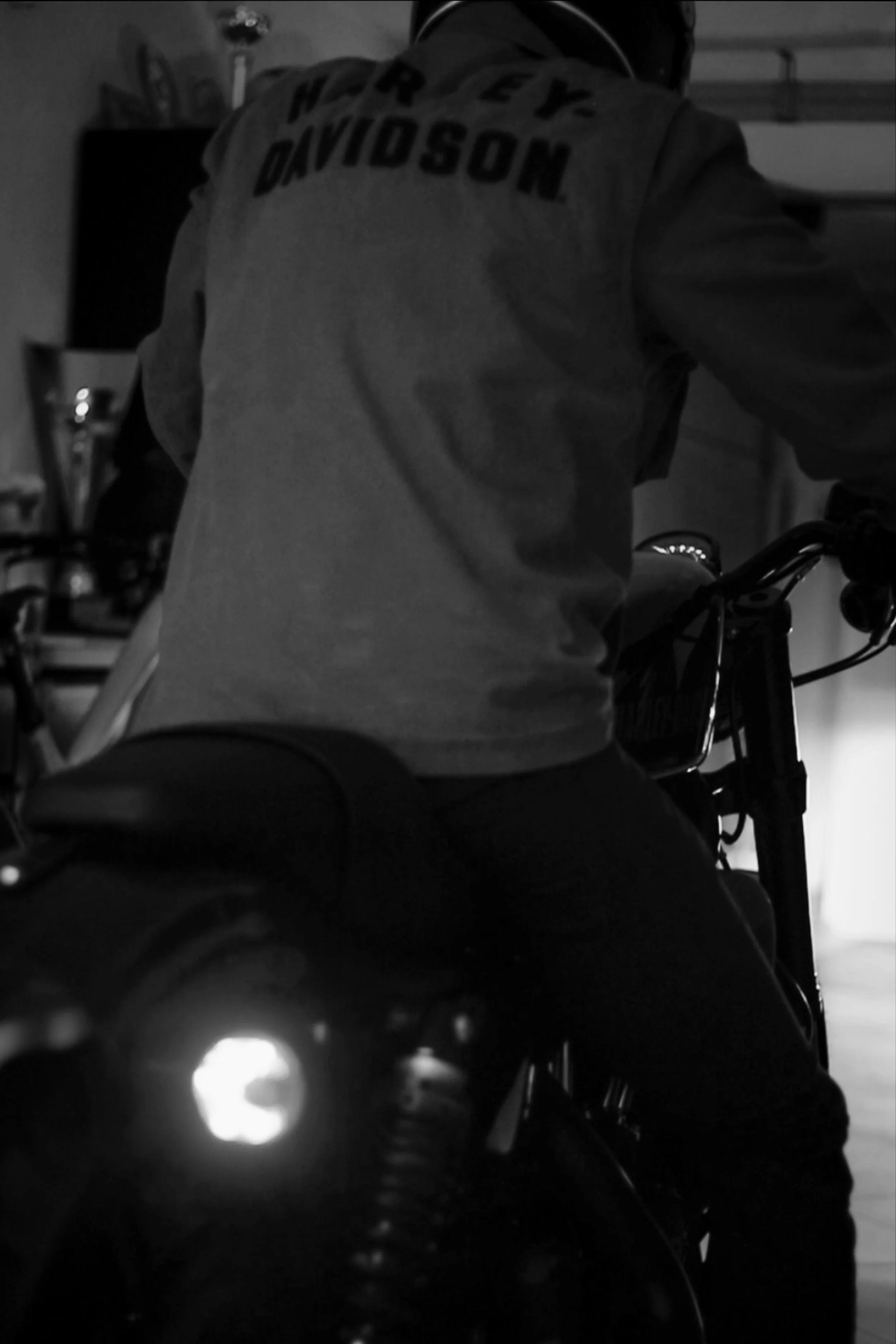 Persona seduta su una motocicletta, vista di schiena, indossa una giacca con 'Harley Davidson' scritto sulla schiena, ambiente interno in ombra.
