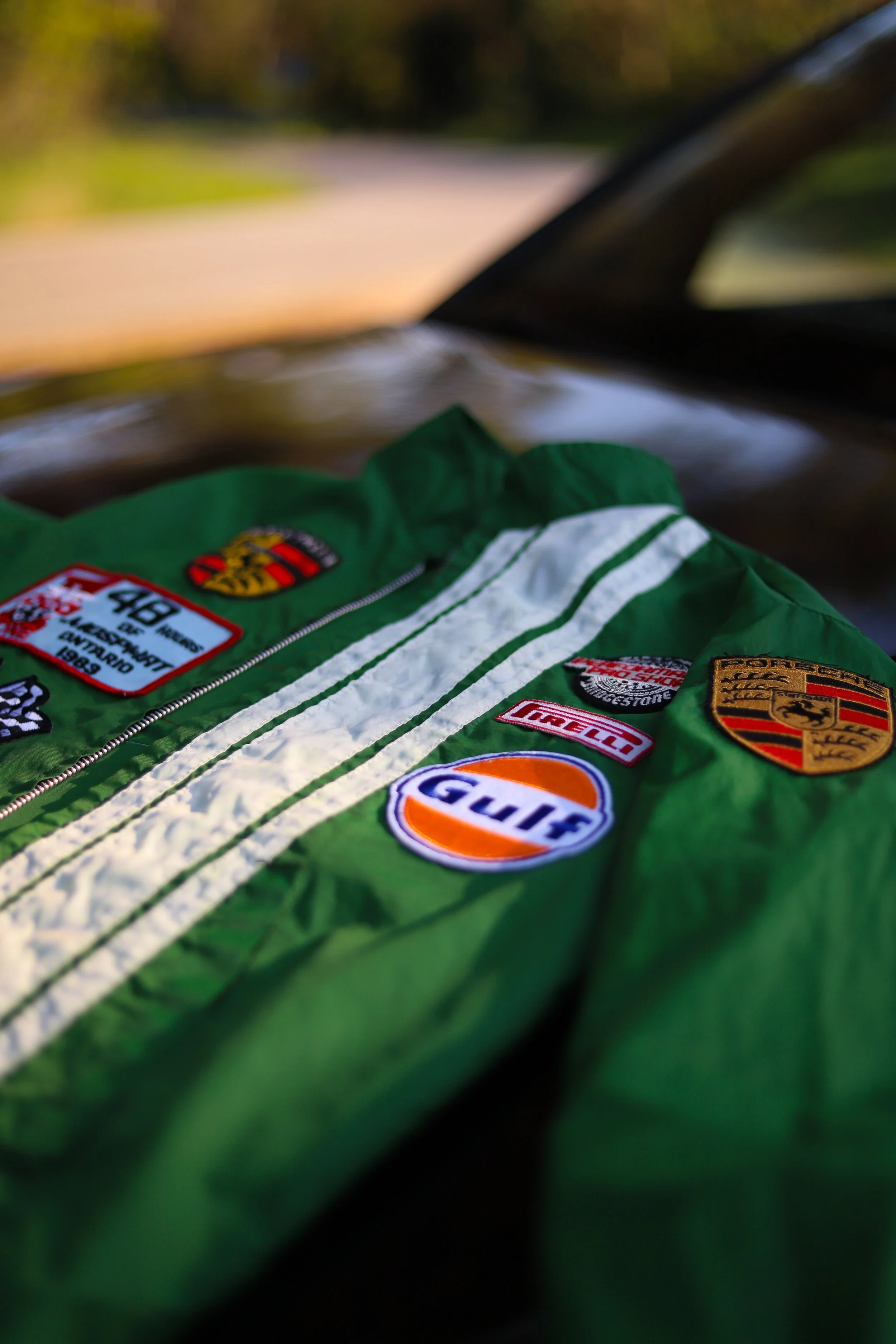 Una giacca da corsa con molte patch e adesivi di marche di automobili da corsa e sportivi, appoggiata su un'auto, con uno sfondo di alberi e cielo.