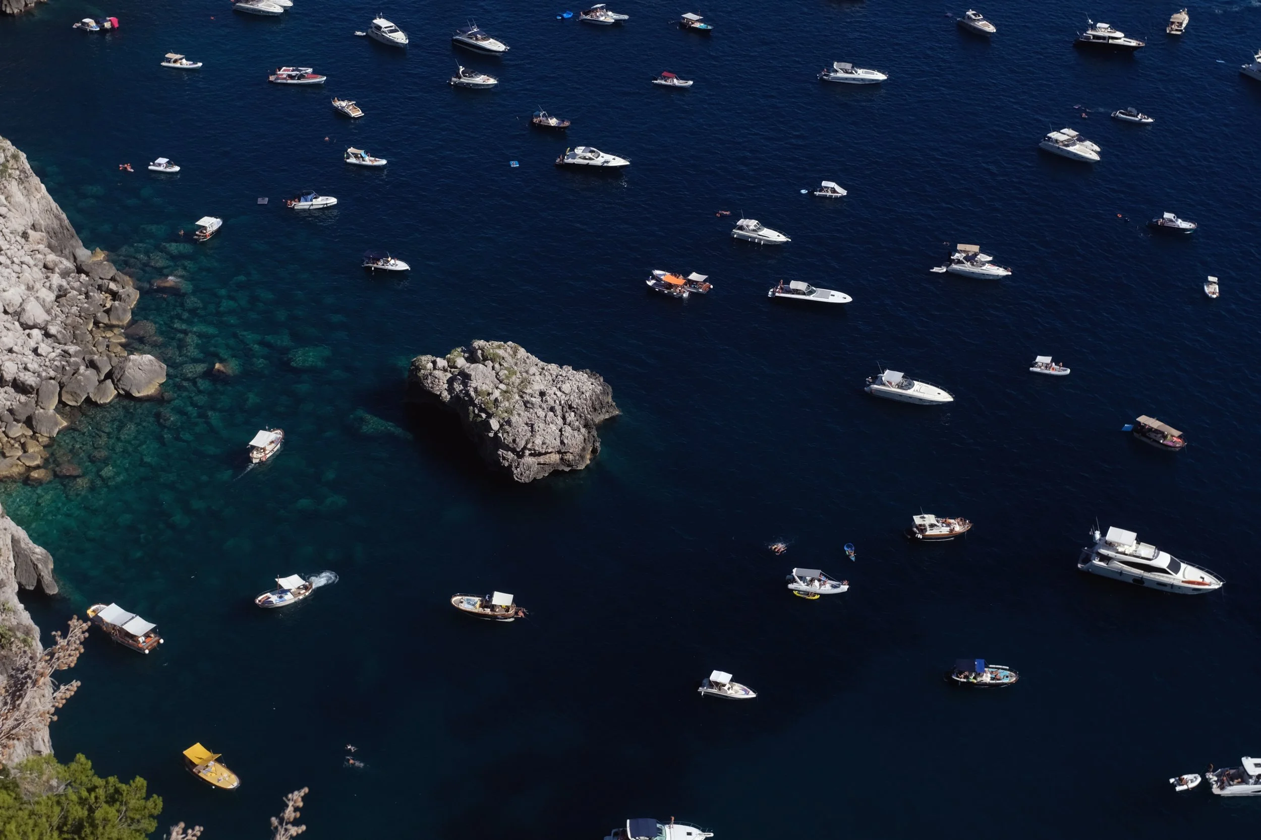 Mare blu con numerose barche ormeggiate vicino a scogli. Acqua trasparente vicino alla riva, con alcune persone che nuotano.